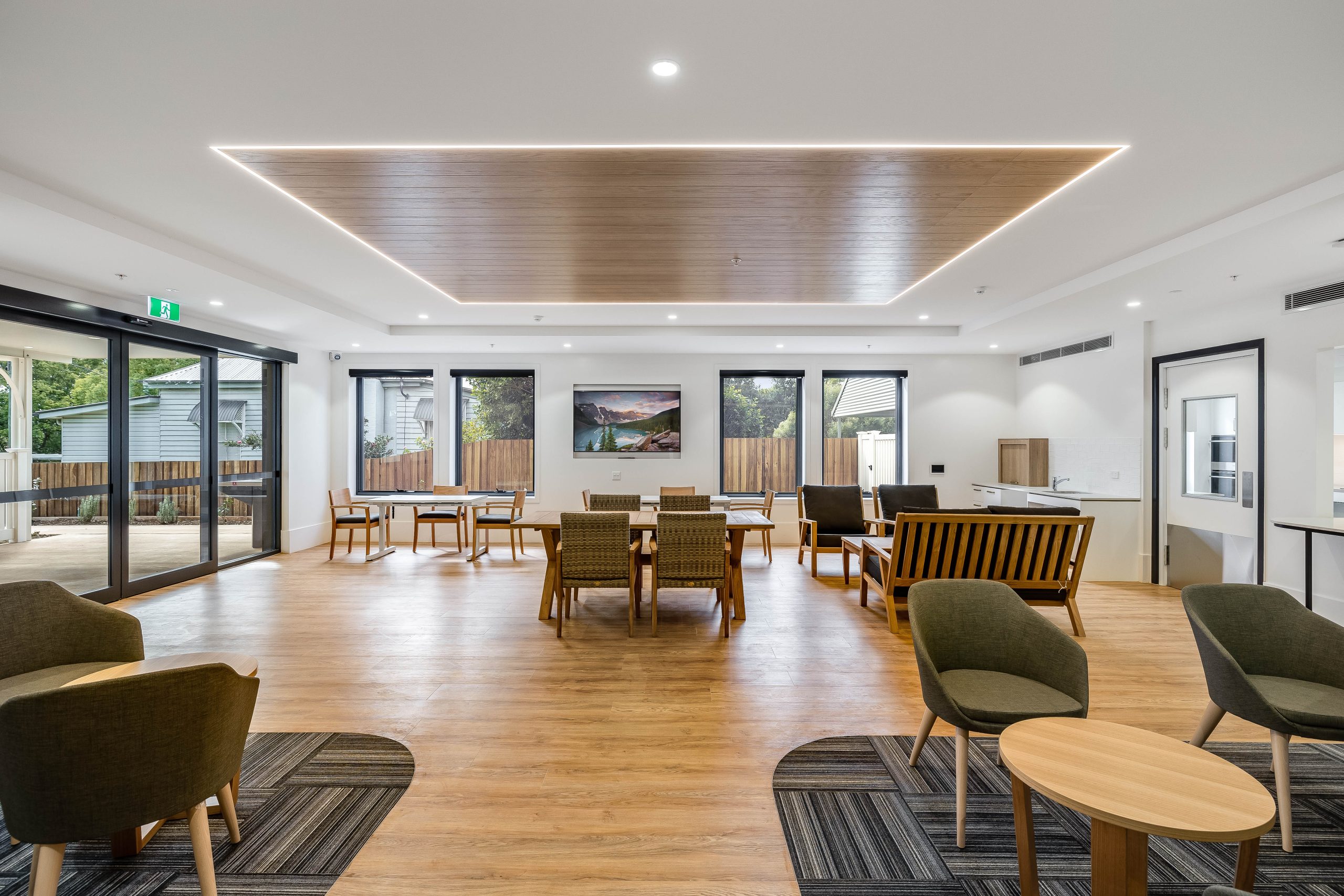 A spacious, modern lounge with wooden floors, green and brown chairs, tables, large windows, and a TV on the wall. Natural light fills the room, and there is access to an outdoor area.