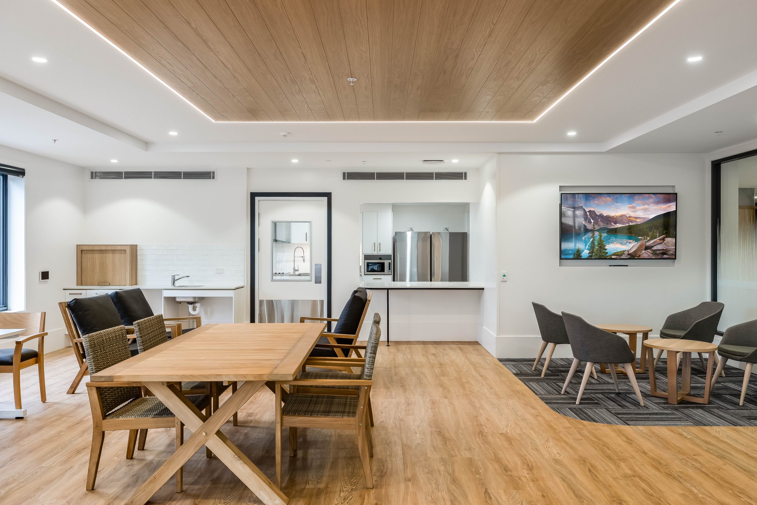 Modern communal room with wooden tables and chairs, a small kitchen area, refrigerator, wall-mounted TV displaying a landscape, and additional seating with armchairs on a rug; wood and white decor.