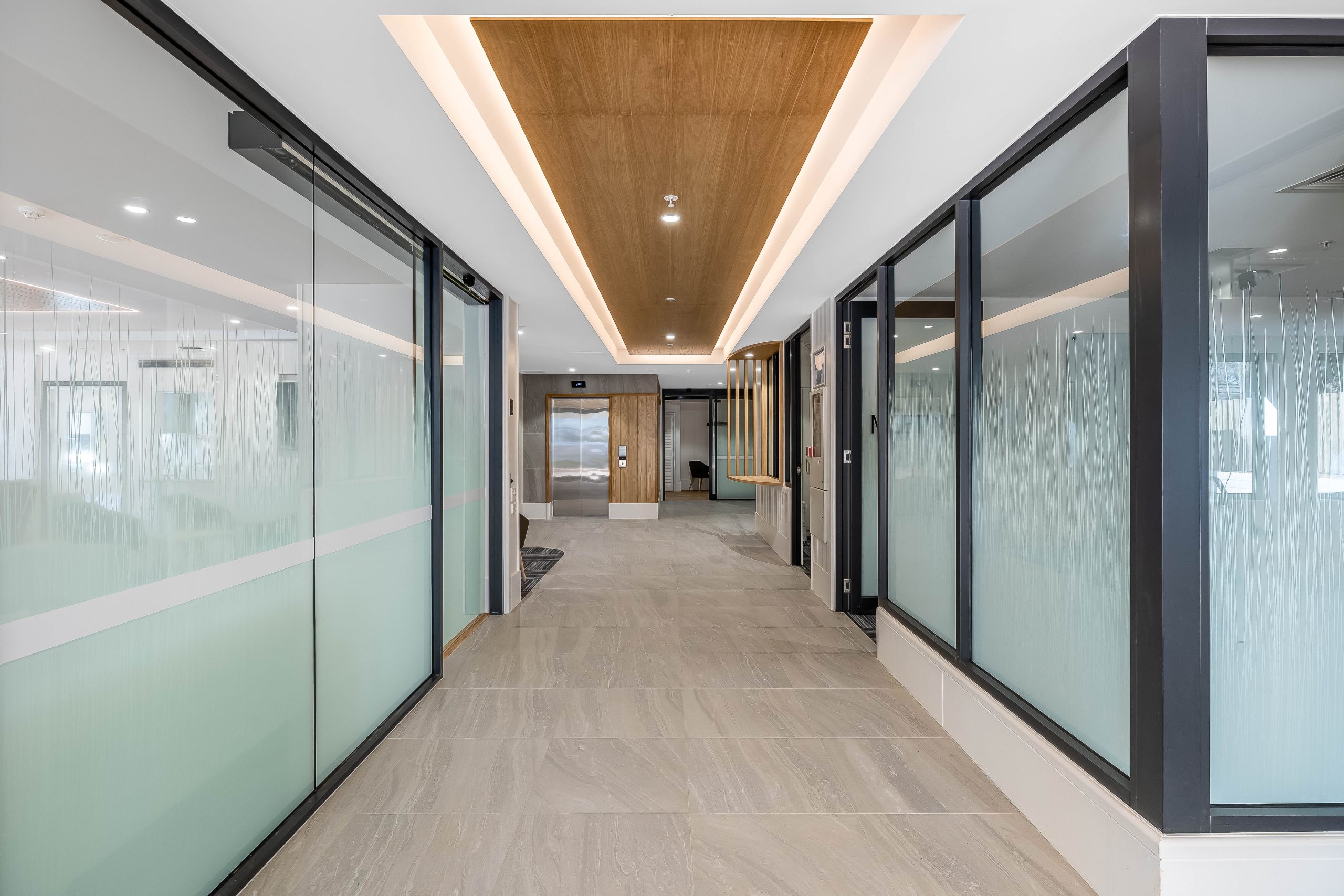 A modern office hallway with frosted glass walls, light-colored tile flooring, wood accents on the ceiling, and an elevator at the end of the corridor.
