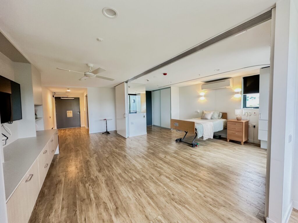 A spacious, modern hospital room with wood-look flooring, a bed near large windows, bedside tables, a wheelchair, TV, and built-in storage along one wall. The room appears bright and clean.