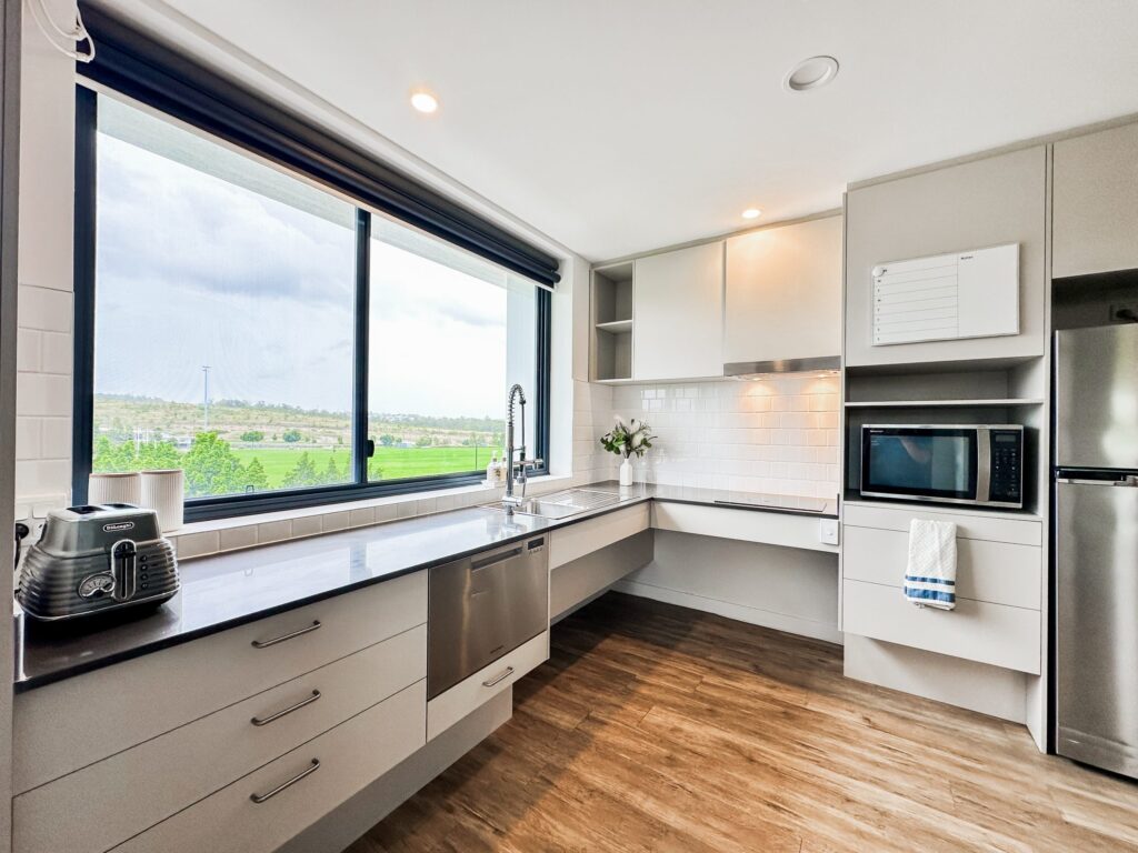 Modern kitchen with light gray cabinets, black countertops, stainless steel appliances, a large window with a view of greenery, wooden floor, and white subway tile backsplash. Toaster and microwave are visible on the counters.