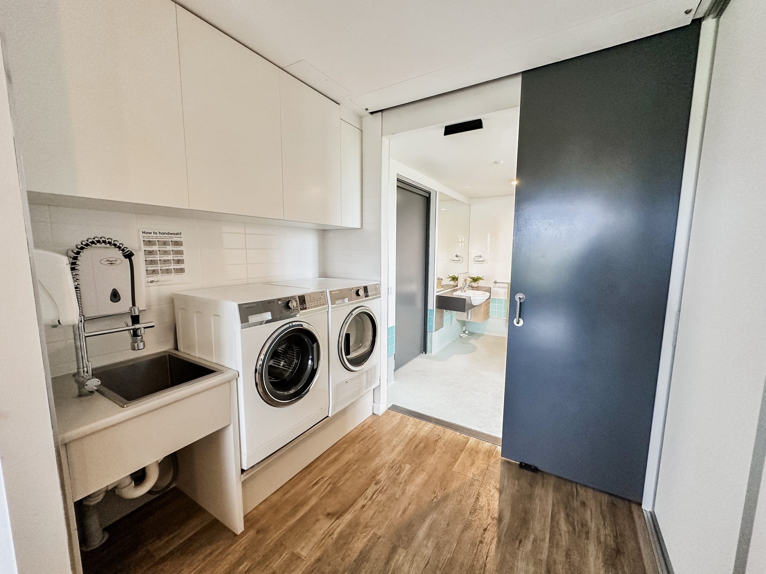 A modern laundry room with a washer, dryer, utility sink, cabinets above, wood flooring, and a sliding dark door leading to a bright bathroom area in the background.