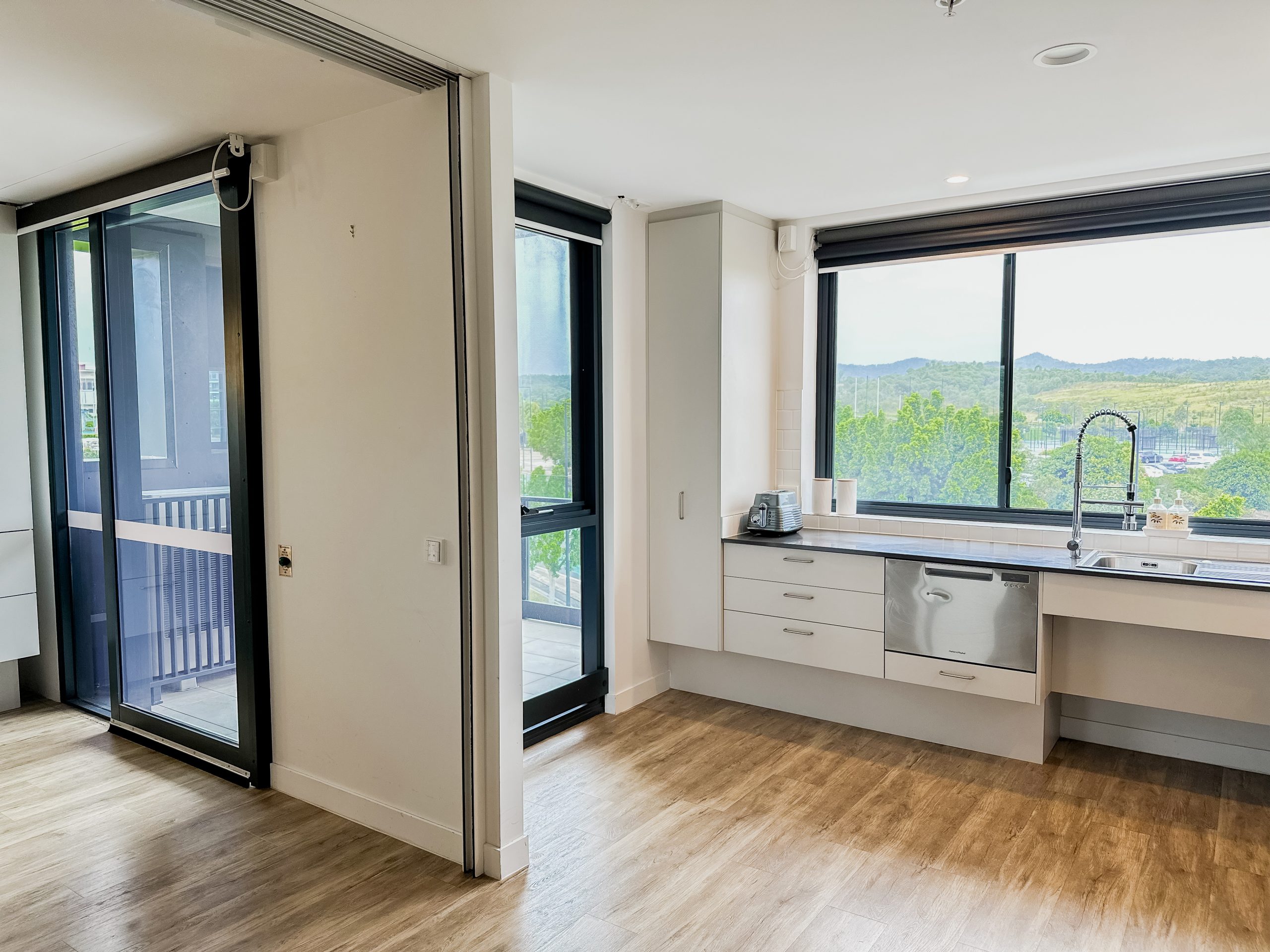 Modern, unfurnished apartment with wood flooring, large windows, and a kitchen area featuring white cabinets, a sink, and a dishwasher. A sliding glass door opens to a balcony with a scenic, green view outside.