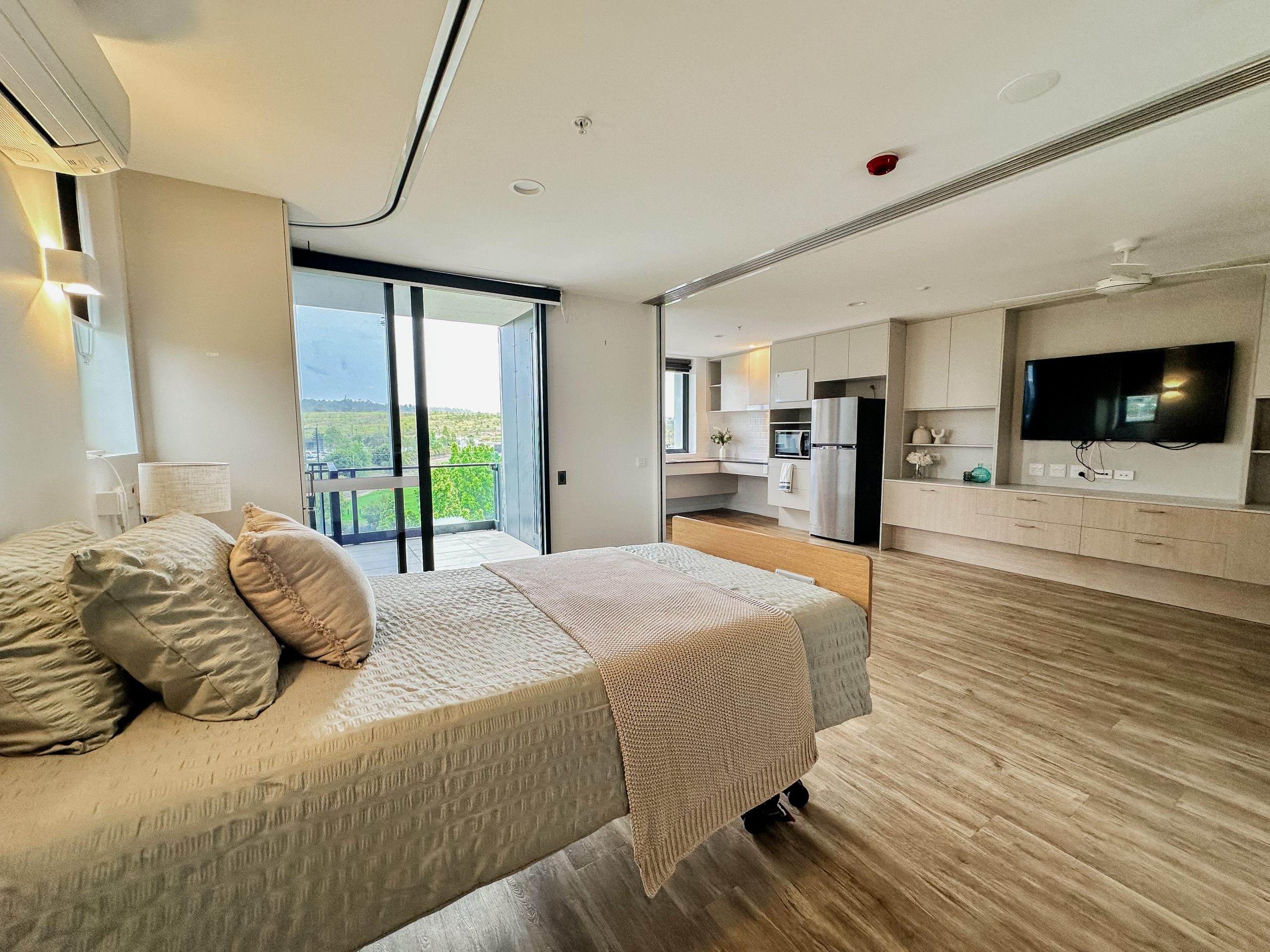 Modern, spacious bedroom with light wood flooring, a neatly made bed, large TV, built-in cabinets, and floor-to-ceiling sliding glass doors opening to a balcony with a green outdoor view.