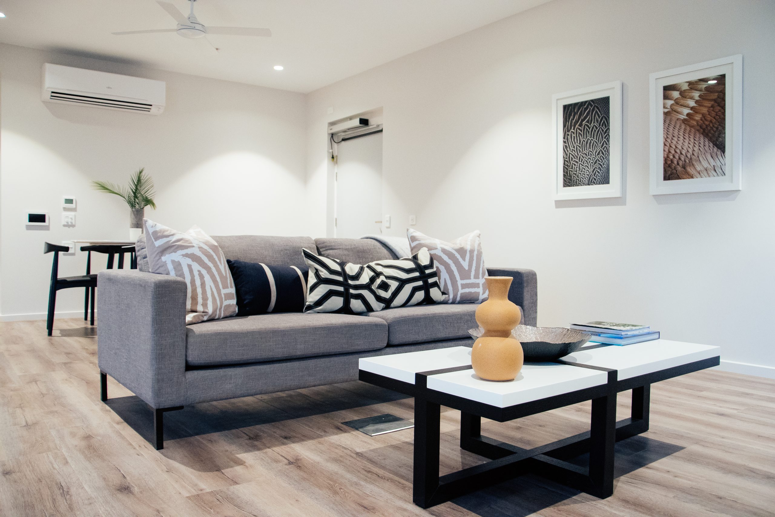 A modern living room with a gray sofa decorated with patterned cushions, a white and black coffee table holding a vase and books, and two framed artworks on the wall. The space has wooden flooring and minimal decor.