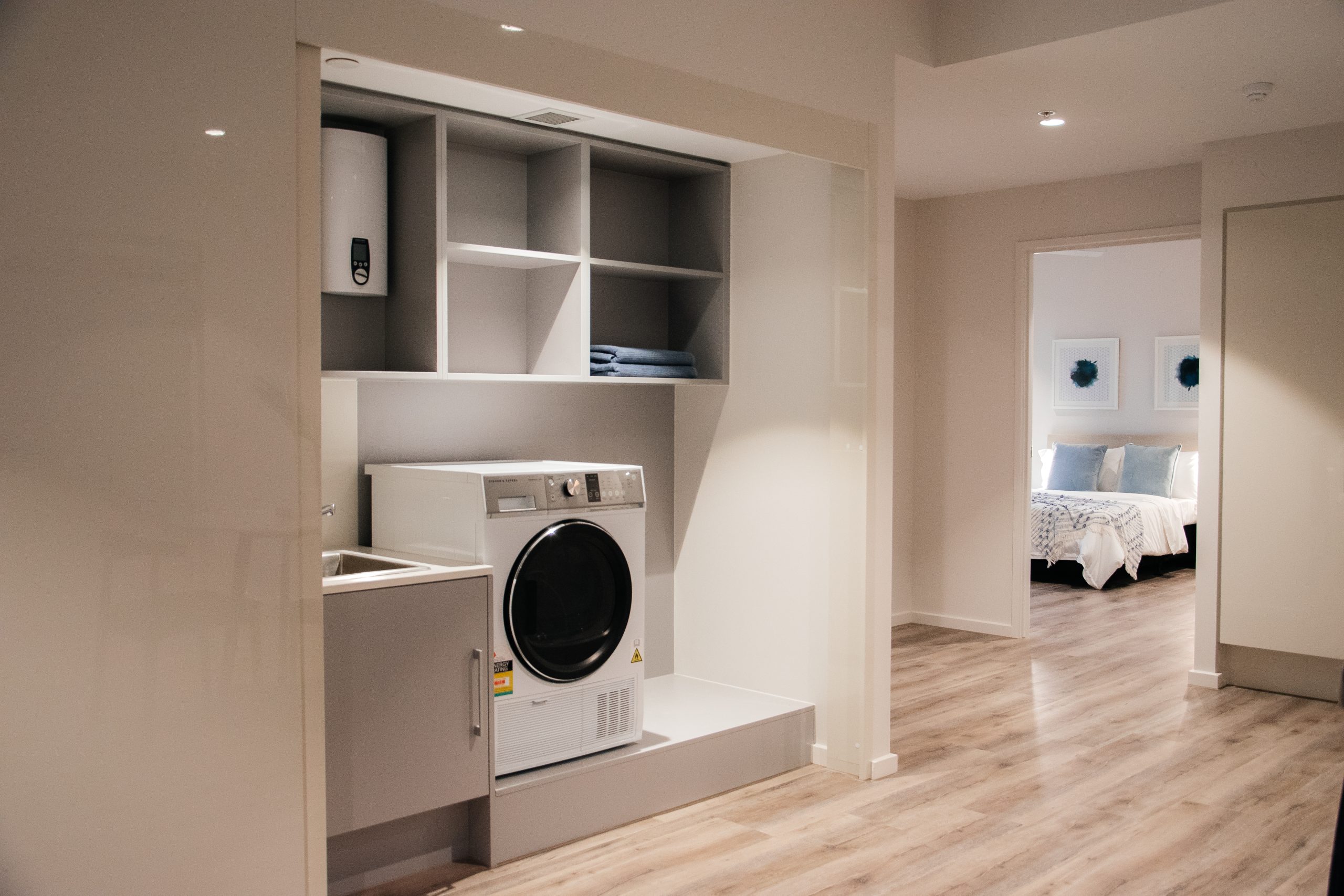 A modern laundry area with a front-loading washing machine, a small sink, open shelves with folded towels, and a water heater, located in a bright room with wood flooring; a bedroom is visible in the background.