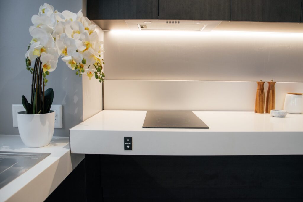 Modern kitchen countertop with a white orchid plant in a pot, an electric cooktop, two wooden salt and pepper shakers, and a small round container, all under under-cabinet lighting.