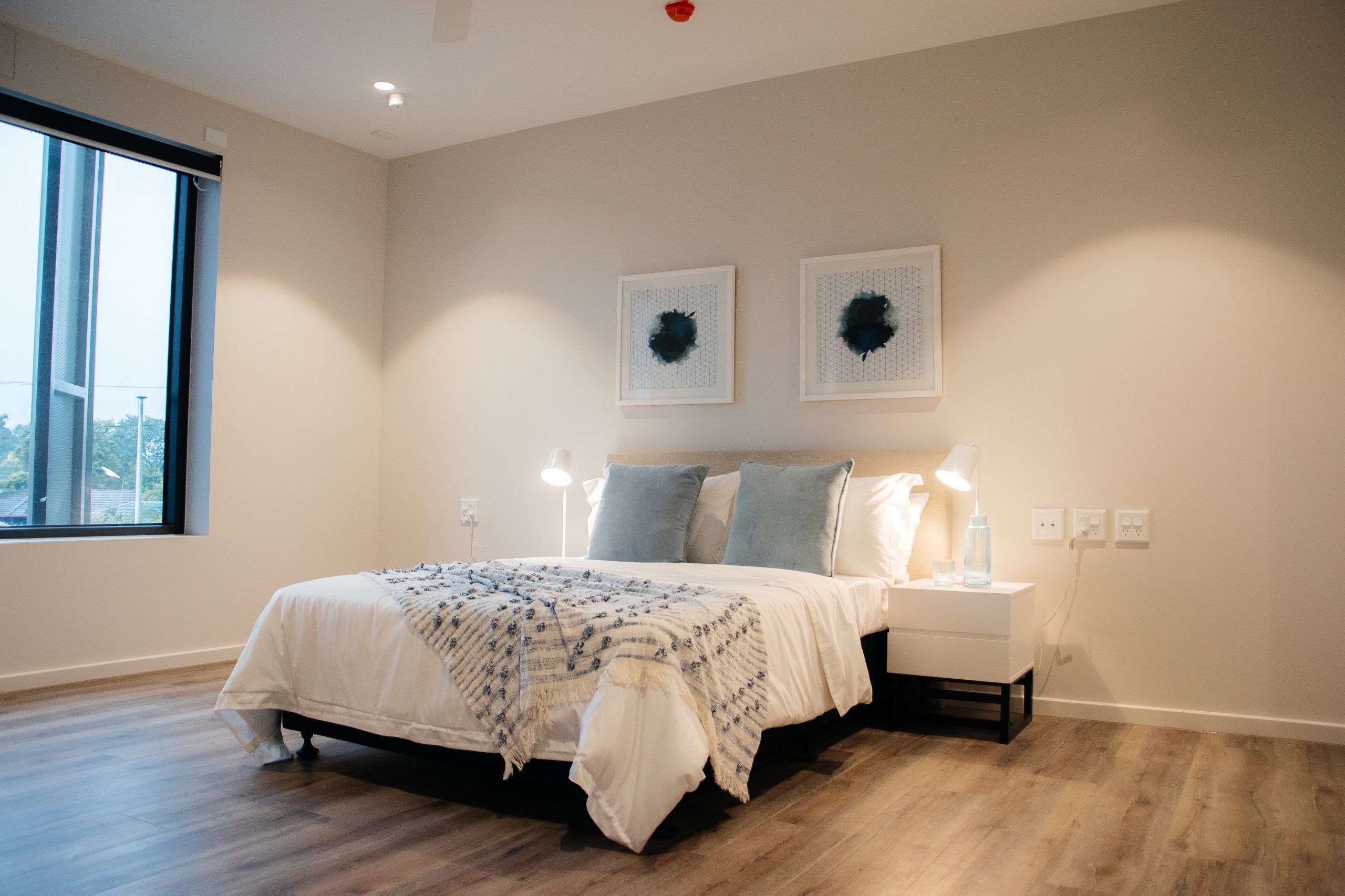 A modern bedroom with a neatly made bed, two gray pillows, white bedding with a striped throw, and two bedside tables with lamps. Two abstract framed artworks hang above the bed. Light wood flooring and a large window are visible.