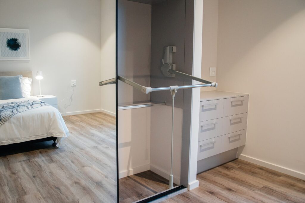 A modern bedroom with light wood flooring, a neatly made bed with white and blue bedding, and an open closet featuring drawers, a mirror, and horizontal hanging rods. The room is softly lit and minimalistic.