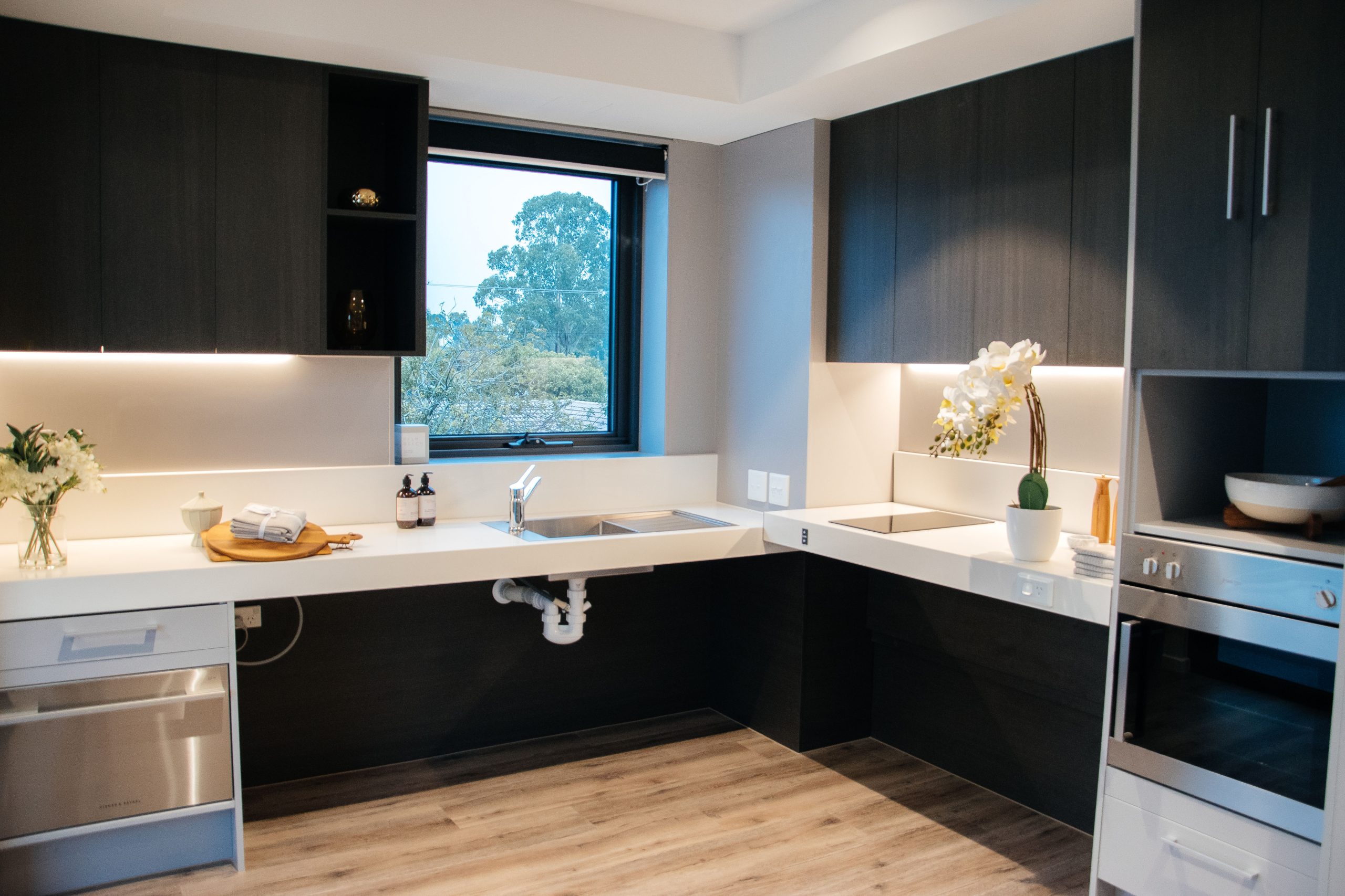 Modern kitchen with dark cabinets, white countertops, under-cabinet lighting, stainless steel sink, oven, wooden floors, a window with trees outside, and decorative flowers on the counters.