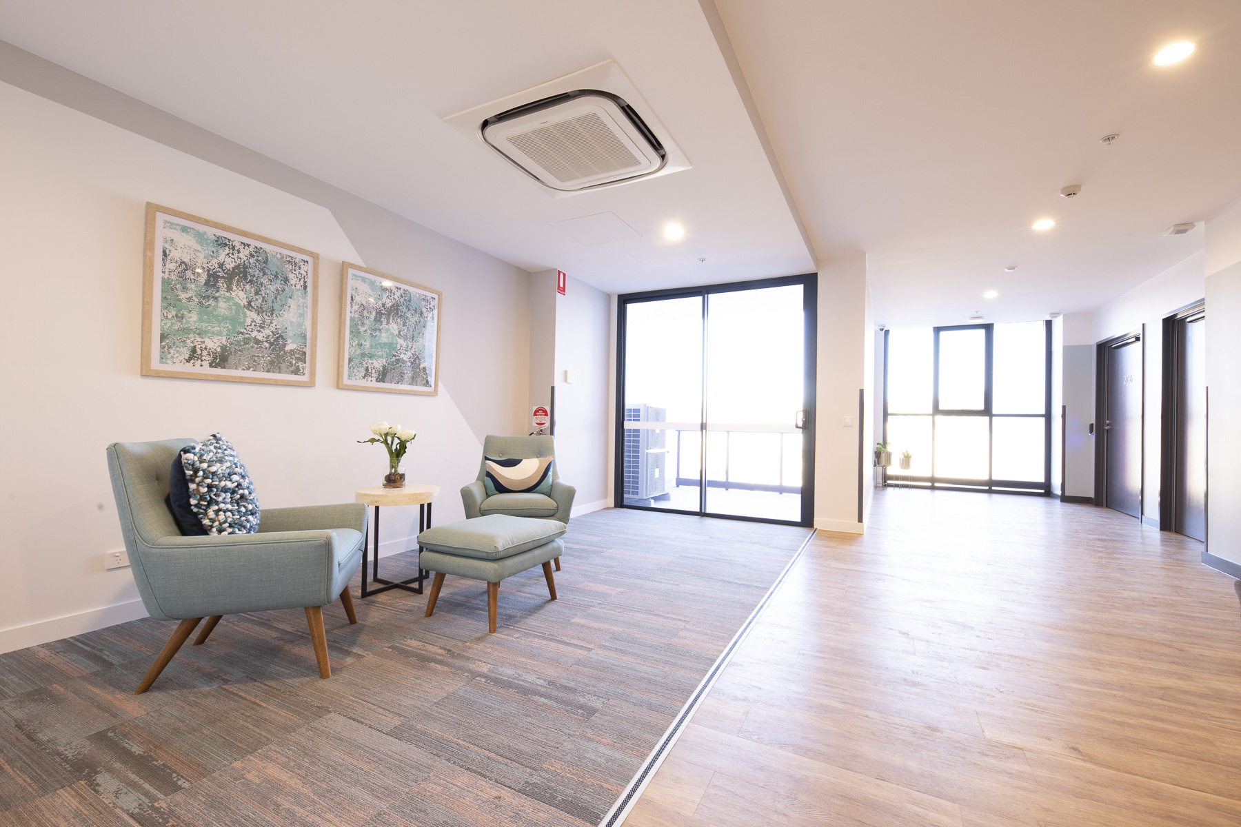 A modern, bright lounge area with two blue armchairs, a small white side table, wall art, and large windows letting in natural light. The space has wood flooring and light-colored walls.
