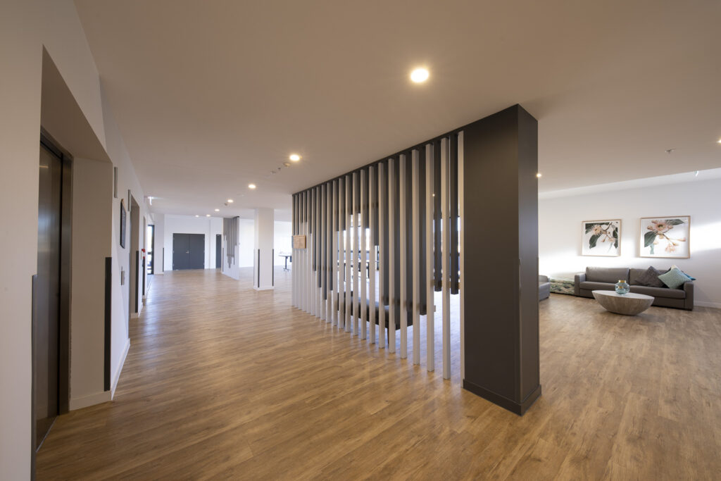 A modern, spacious hallway with wooden floors, vertical slat divider, recessed lighting, and a seating area with a couch, coffee table, and framed artwork on the wall in the background.