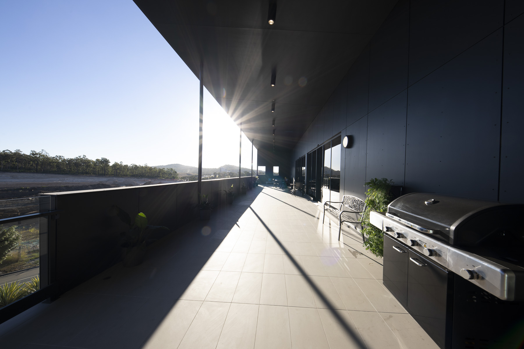 A modern, spacious balcony with a sleek black ceiling, glass railing, grill, and outdoor seating. Sunlight creates long shadows across the light-tiled floor, with a scenic view of hills in the background.