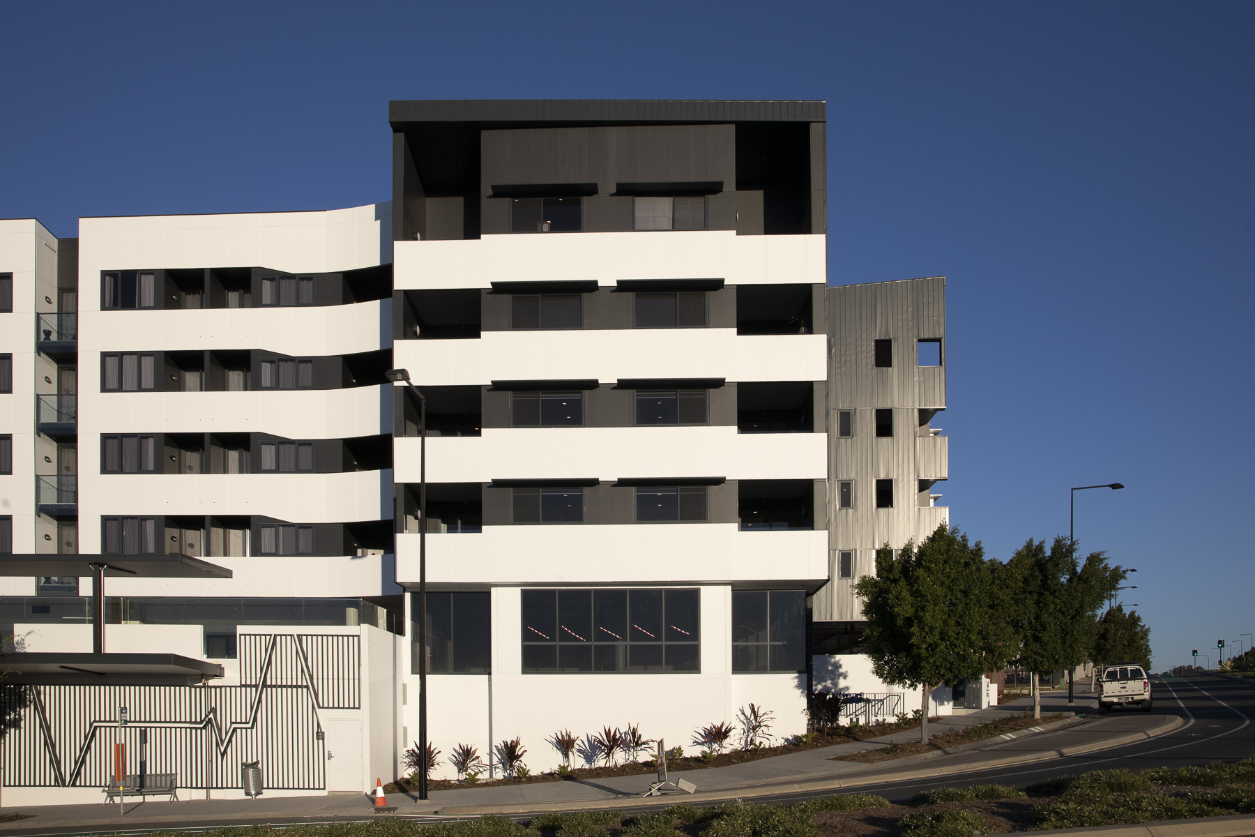 A modern multi-story apartment building with black and white geometric design, large windows, and balconies, situated next to a street with trees and streetlights under a clear blue sky.