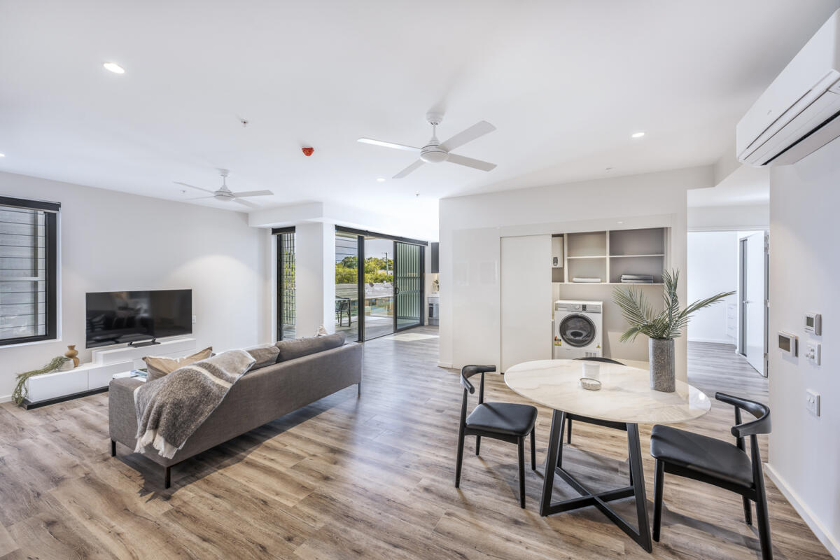 Modern open-plan living area with a gray sofa, TV, round dining table, two chairs, ceiling fans, wood flooring, and large windows offering an outdoor view. A laundry area with a washing machine is visible in the corner.