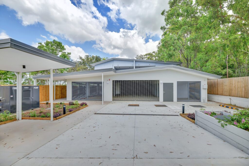 Modern white building with a metal garage door, surrounded by greenery and a wooden fence, with a wide concrete driveway and landscaped garden beds in the foreground under a partly cloudy sky.