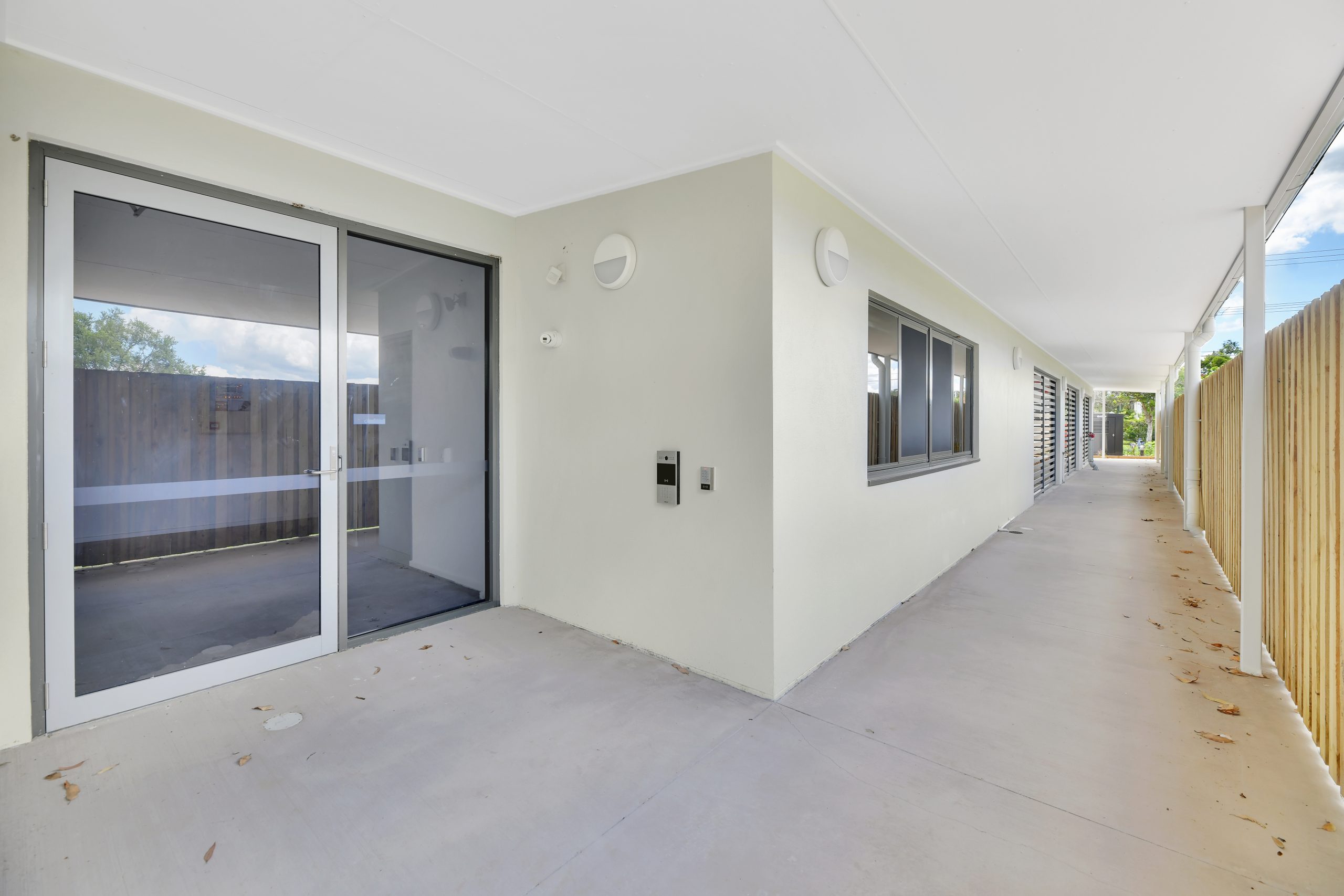 A covered outdoor walkway beside a modern building with large windows, a glass door, and beige walls; the walkway is bordered by a wooden fence and scattered with a few dry leaves.