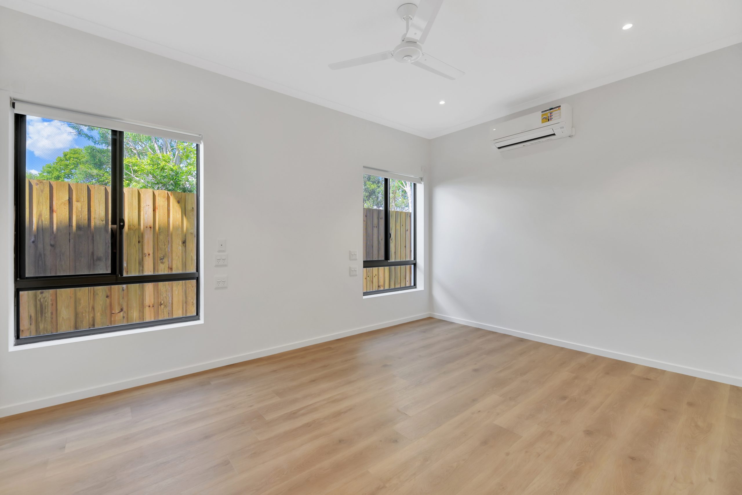 A bright, empty room with light wood flooring, white walls, two windows with wooden fencing outside, a ceiling fan, and a wall-mounted air conditioning unit.