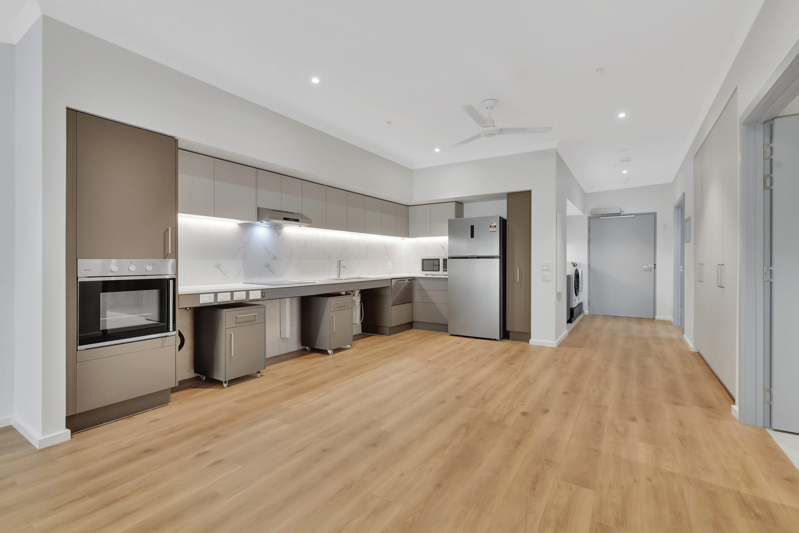 Modern, spacious kitchen with wood flooring, beige and gray cabinets, built-in oven, stovetop, stainless steel refrigerator, and white marble backsplash. Bright lighting and open layout with adjoining hallway.