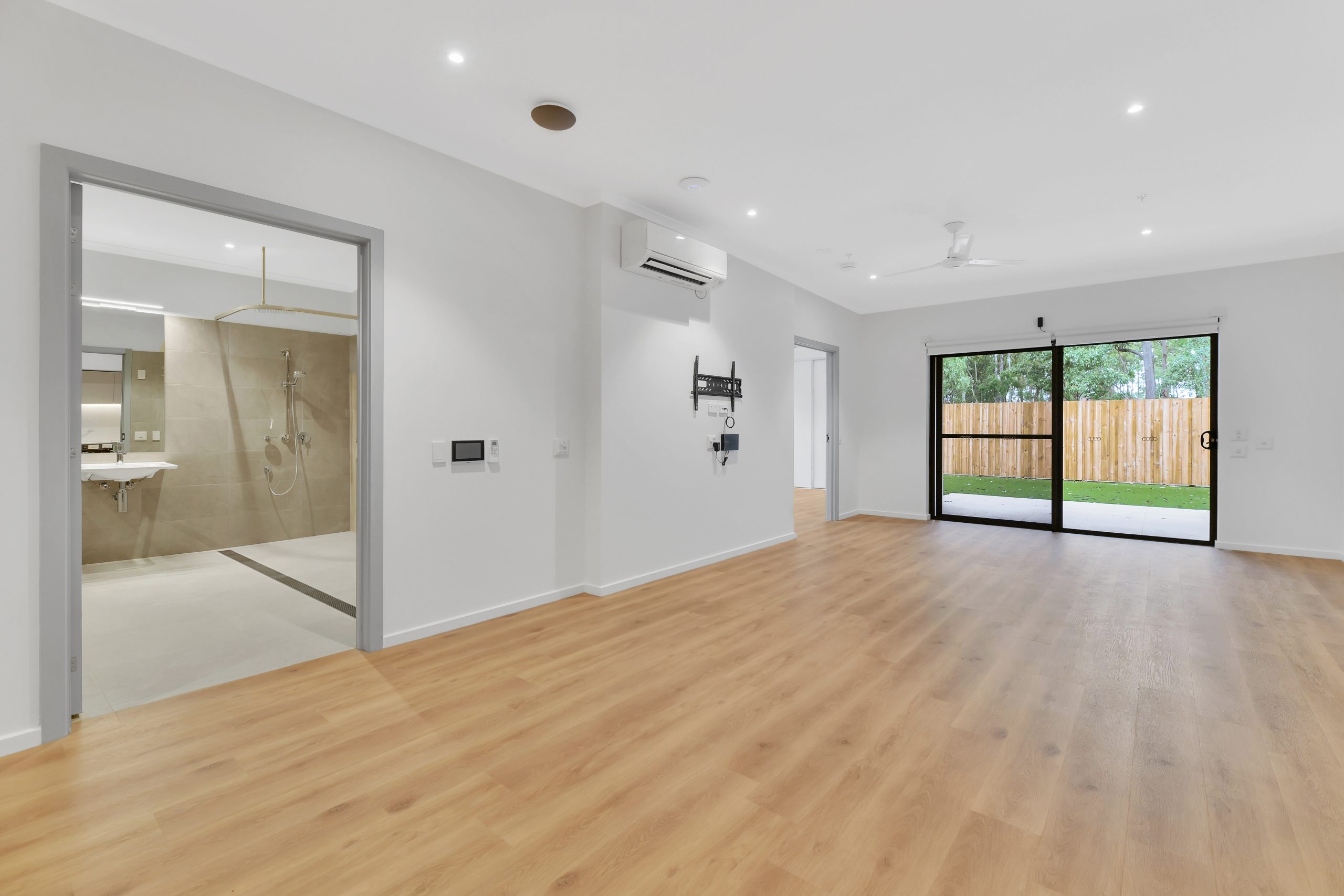 Modern, unfurnished living space with light wood flooring, white walls, recessed lights, ceiling fan, and air conditioning. Visible bathroom on the left and large glass sliding doors leading to a fenced backyard on the right.