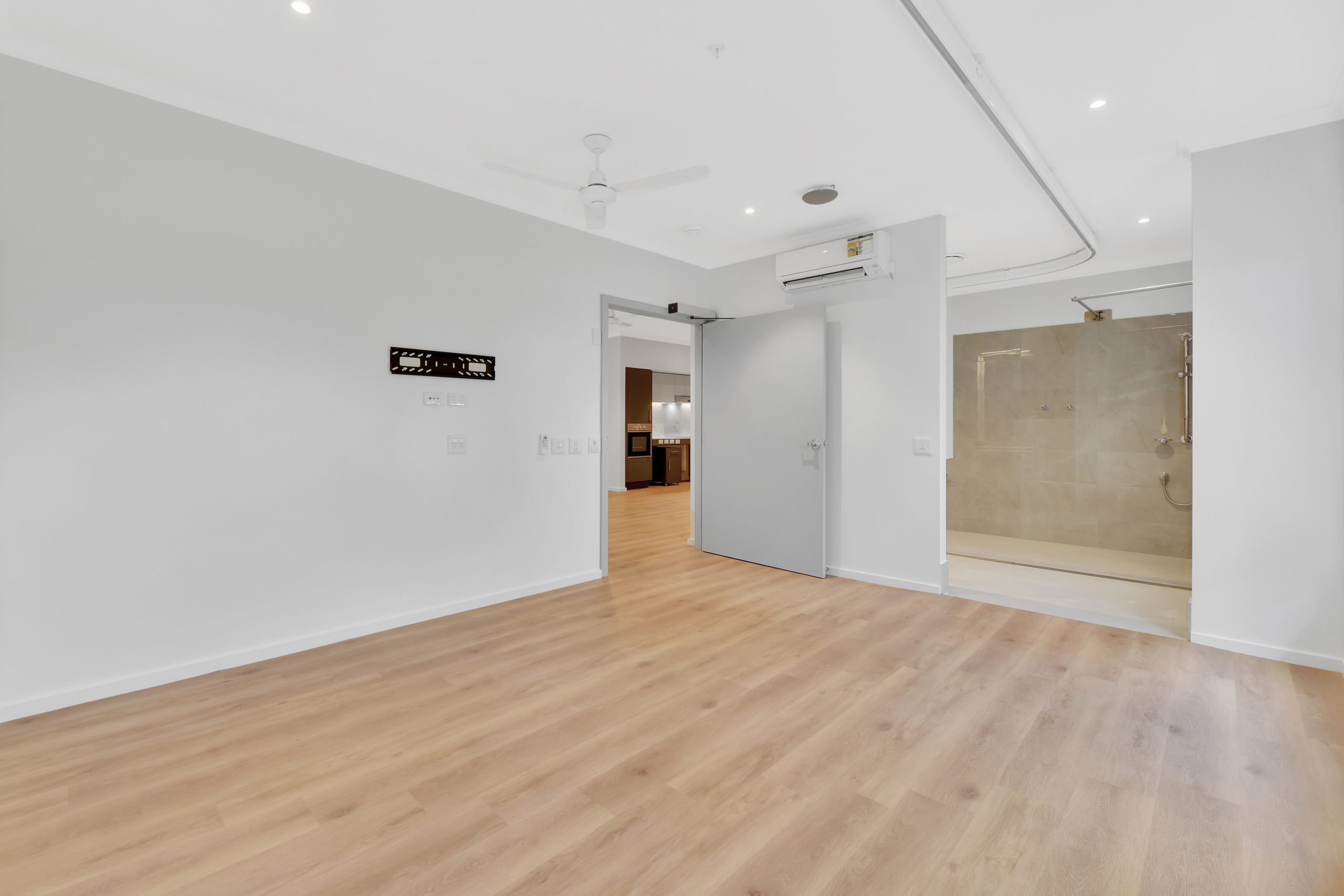 A spacious, empty room with light wooden floors, white walls, a ceiling fan, and an air conditioning unit. An open door reveals a tiled bathroom with a walk-in shower. Recessed ceiling lights brighten the space.