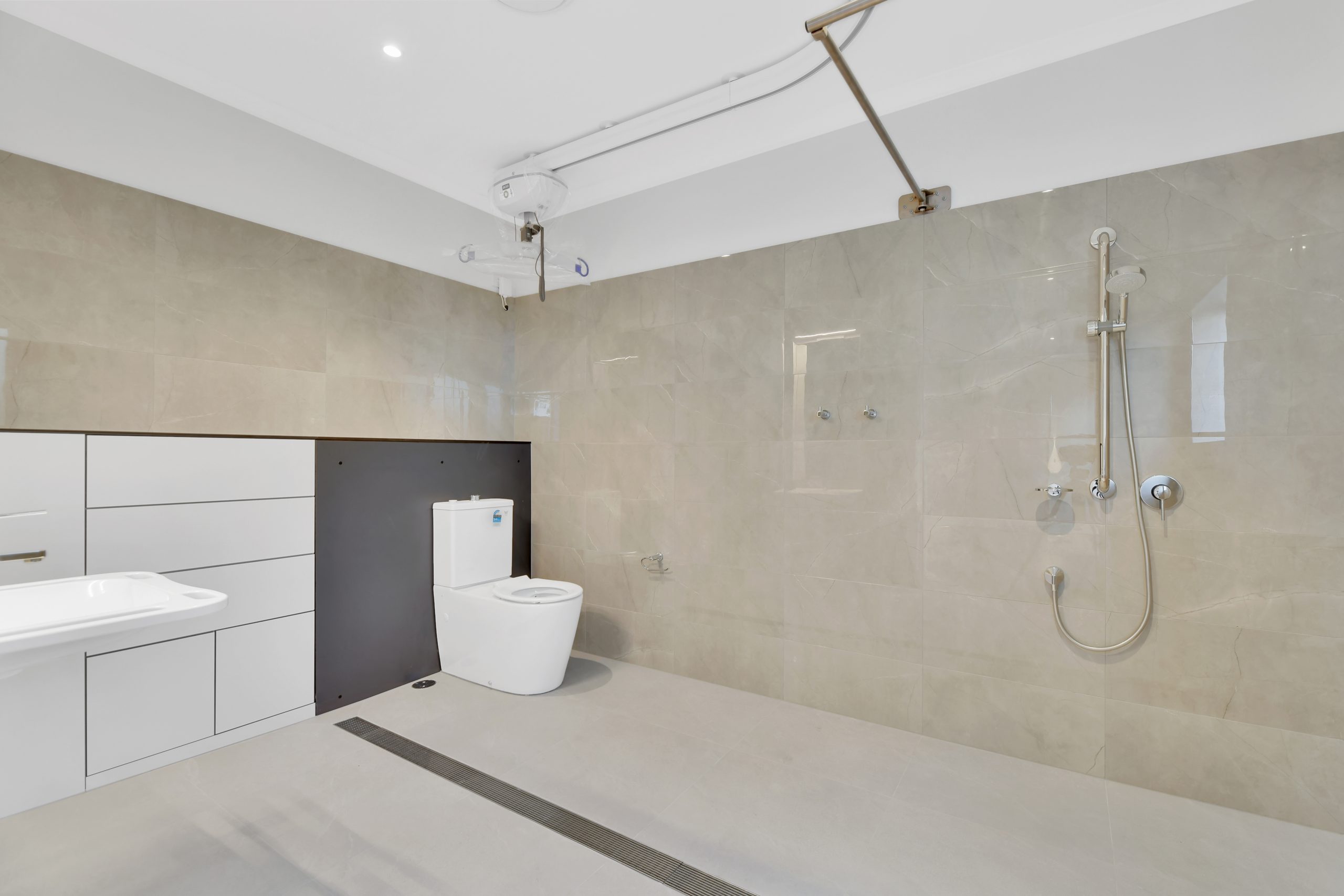 Modern accessible bathroom with a wall-mounted toilet, open shower with handheld showerhead and grab rails, a white sink with cabinetry, and beige tiled walls and floor.