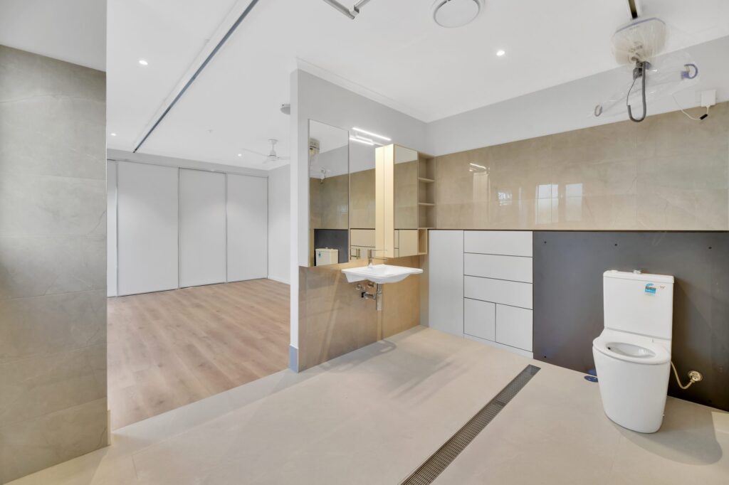 Modern, spacious accessible bathroom with beige and white tiles, wall-mounted sink, grab bars, open shelving, toilet, a ceiling hoist, and a large open shower area. Wooden flooring is visible in the adjacent room.