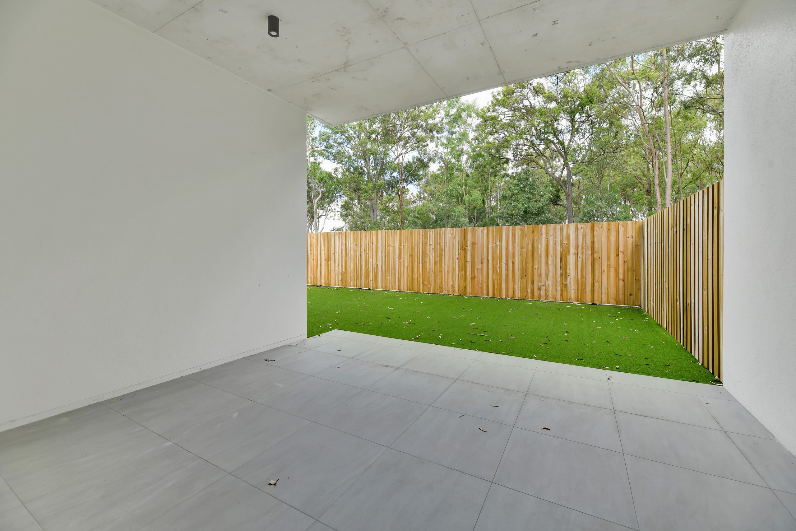 A covered patio with gray tiles opens to a backyard with artificial grass, bordered by a wooden privacy fence and surrounded by tall green trees.