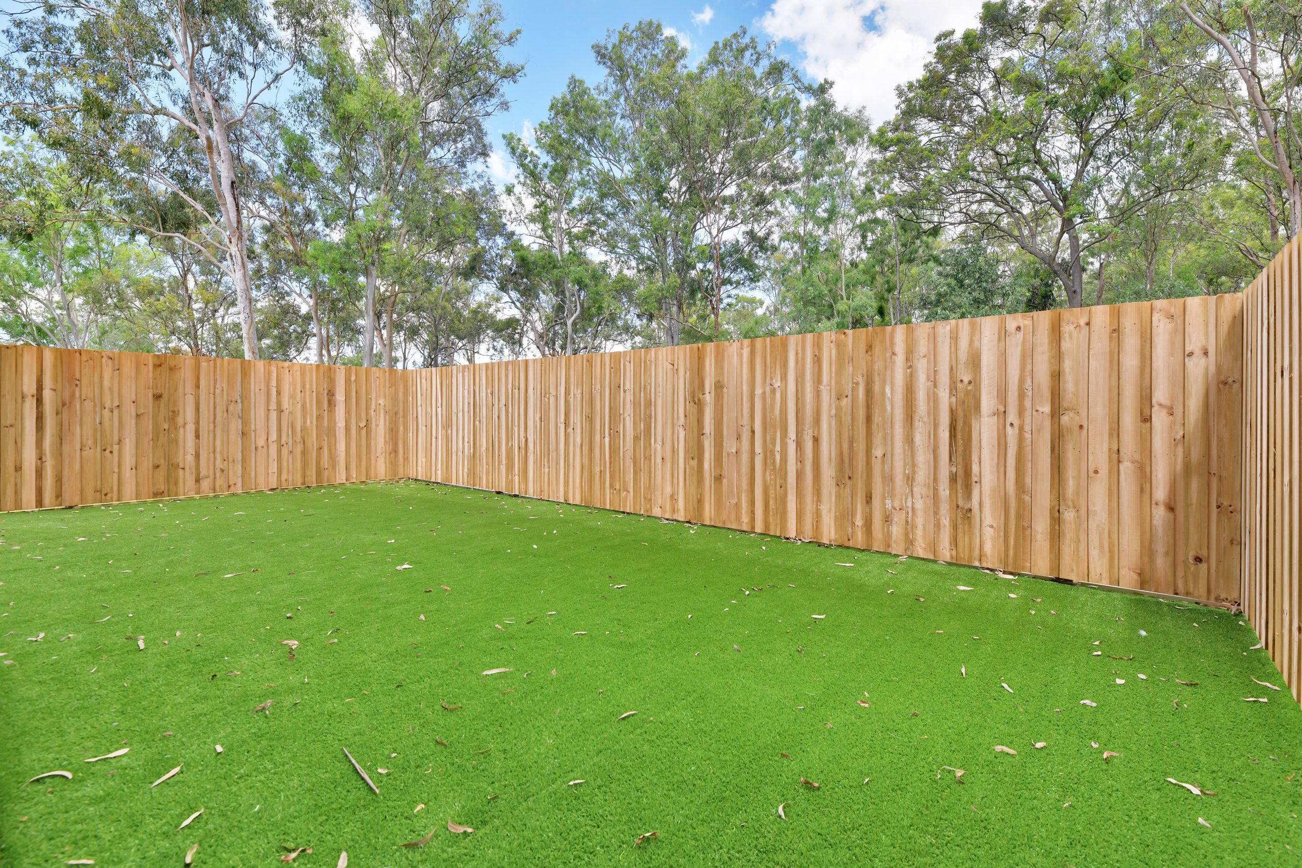 A backyard with bright green artificial grass, surrounded by a tall wooden fence, with trees and a partly cloudy sky visible in the background.