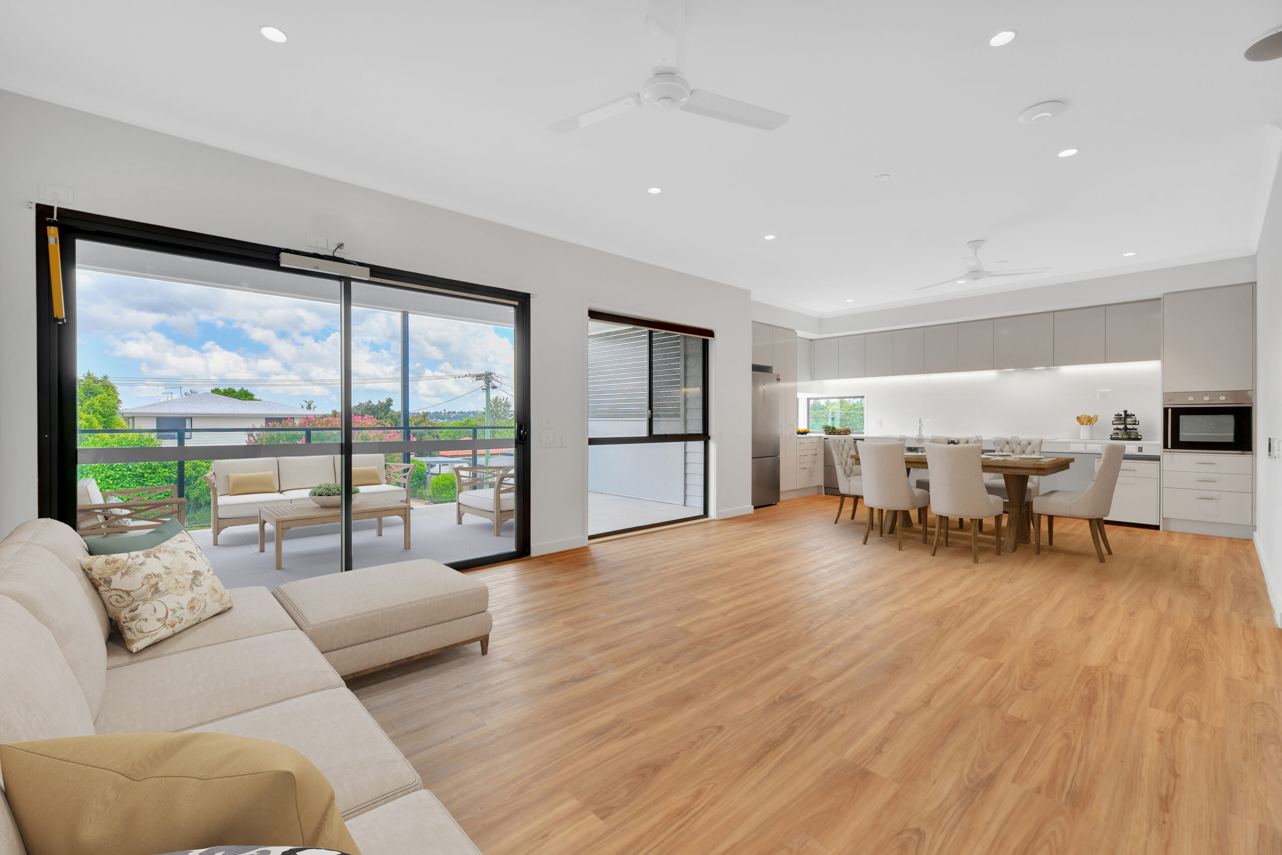 Open-plan living area with light wood flooring, a beige sectional sofa, sliding glass doors to a balcony with outdoor seating, and a modern kitchen with white cabinets and a dining table for six.