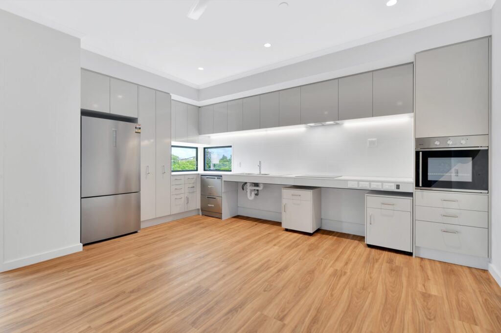 Modern kitchen with light wood flooring, gray cabinets, stainless steel refrigerator, built-in oven, white countertops, and a large window providing natural light above the sink area.