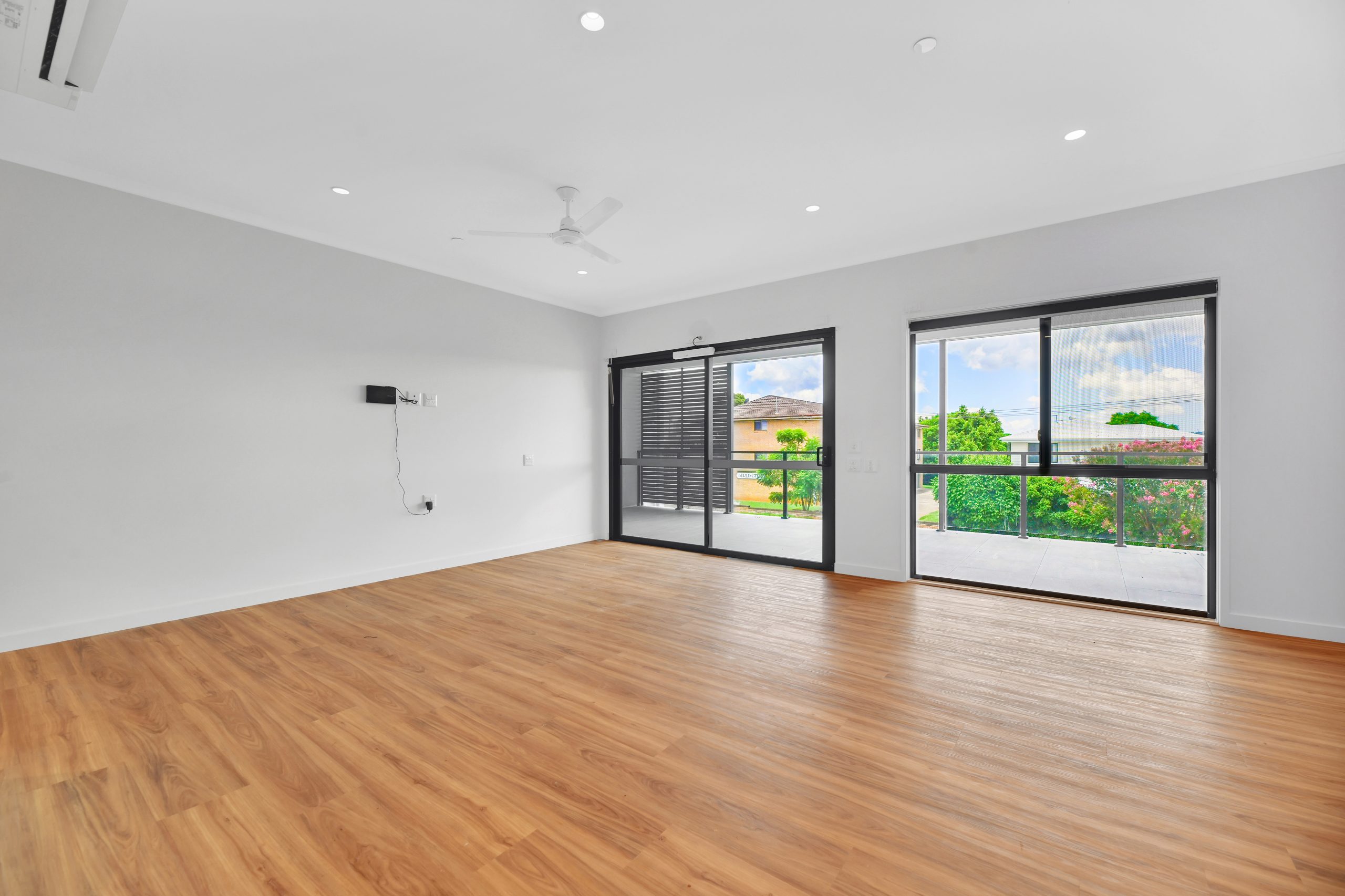 A spacious, empty room with light wood flooring, white walls, a ceiling fan, and large windows letting in natural light. The windows offer a view of greenery and a neighboring house outside.