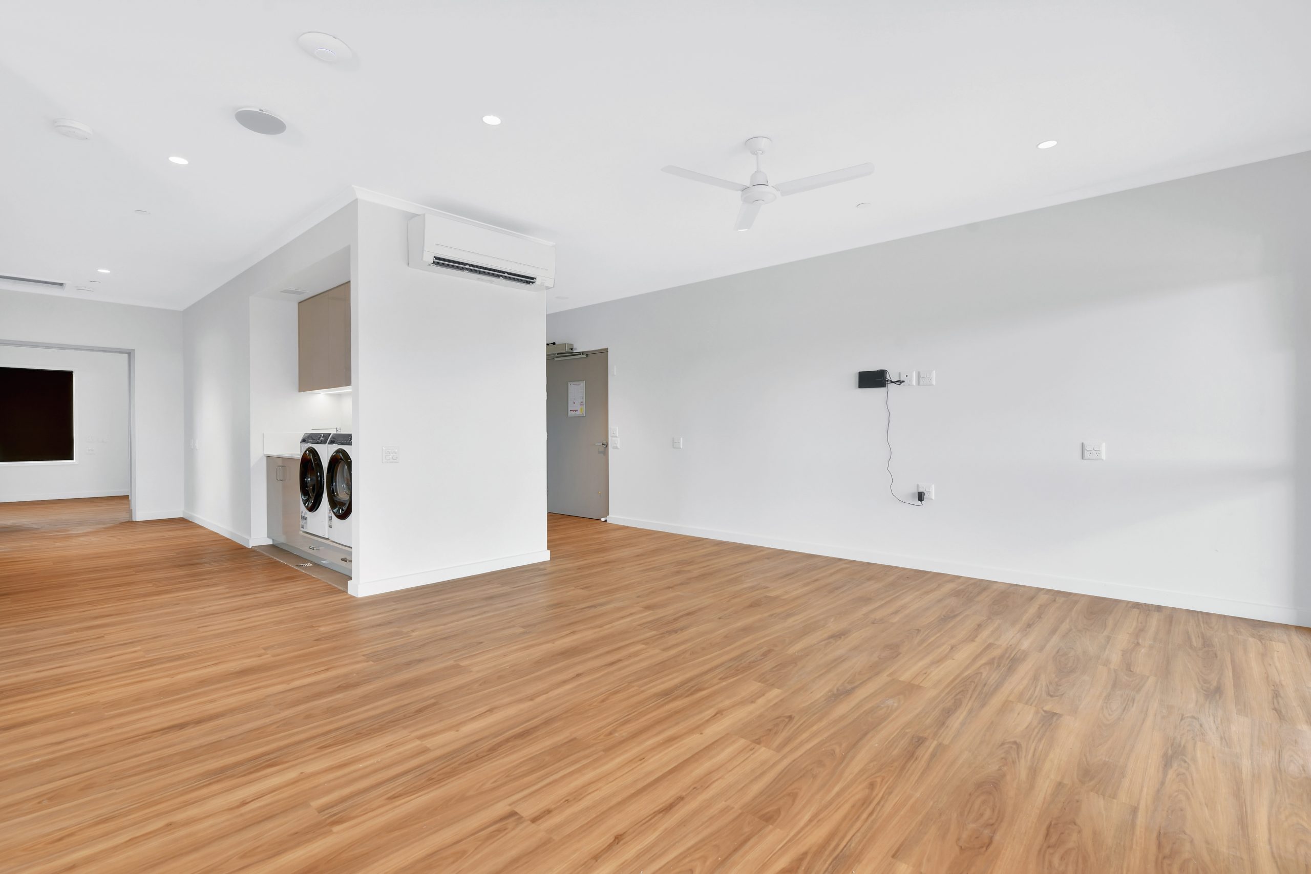 A spacious, unfurnished room with light wood floors, white walls, a ceiling fan, wall-mounted air conditioner, a laundry nook with washer and dryer, and a door in the background.
