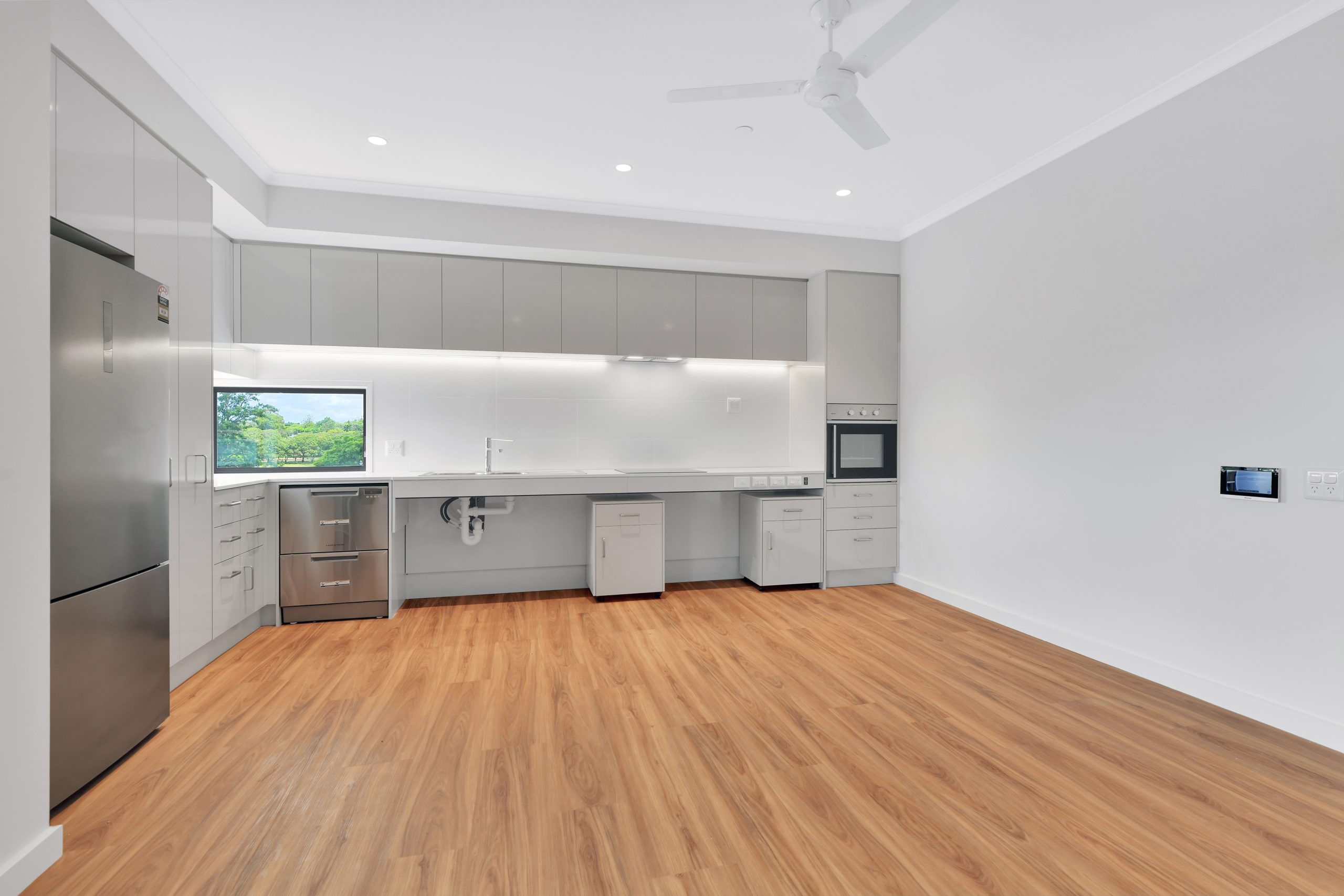Modern, spacious kitchen with light wood flooring, grey cabinets, stainless steel fridge and oven, white countertops, under-cabinet lighting, a large window, and a ceiling fan. Walls and ceiling are white.
