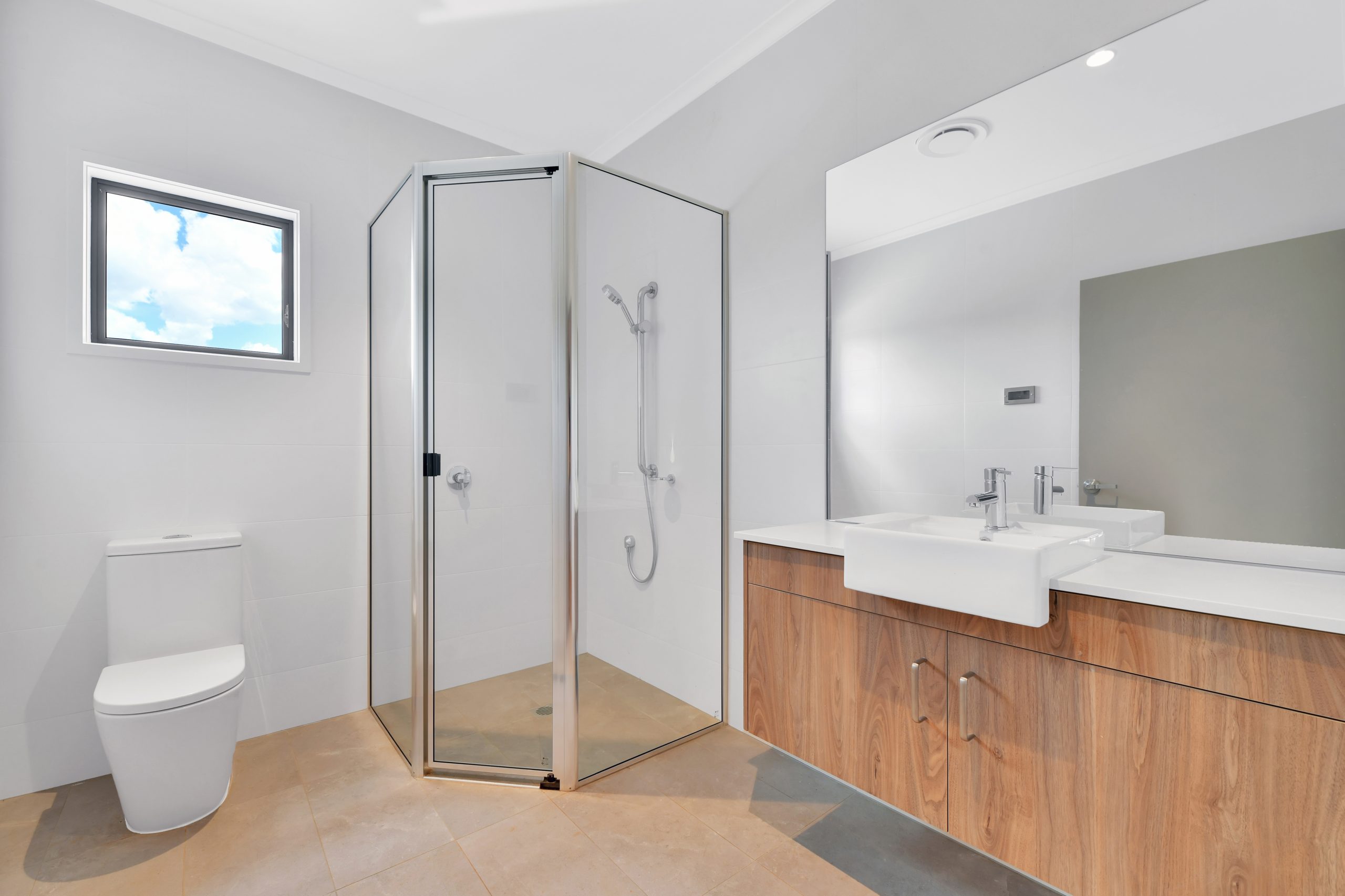Modern bathroom with a glass-enclosed corner shower, a white toilet, and a wooden vanity topped with a rectangular sink and large mirror. A small window lets in natural light.
