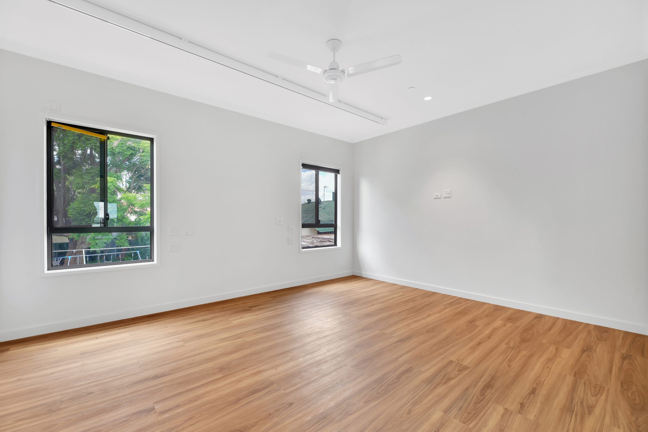Spacious empty room with light wood flooring, white walls, two windows letting in natural light, a white ceiling fan, and minimalistic design.