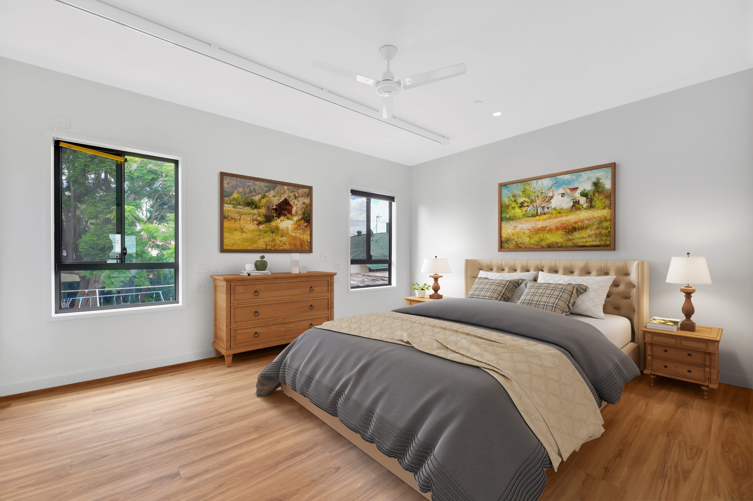 A bright bedroom with wooden floors, a double bed with gray and beige bedding, two nightstands with lamps, a wooden dresser, paintings on the walls, and large windows letting in natural light.