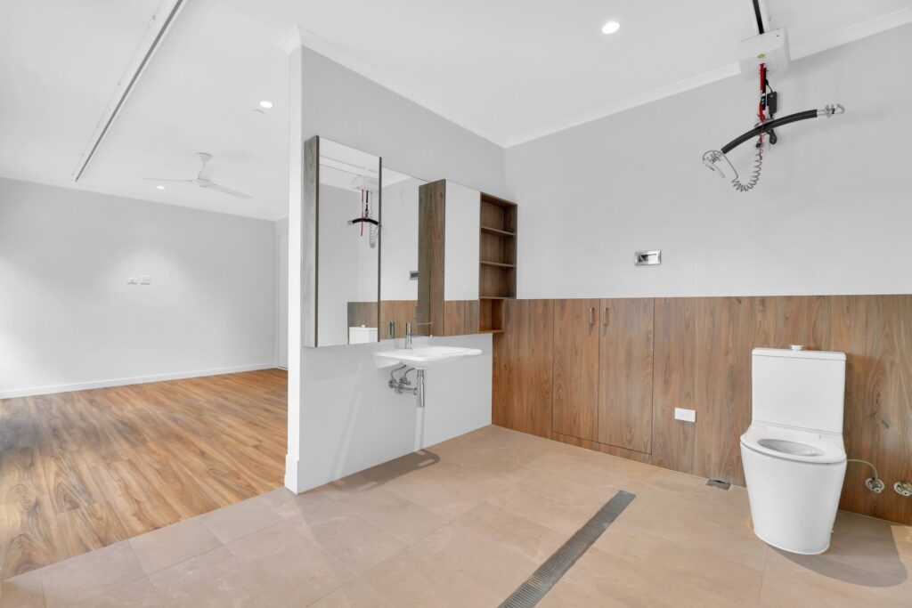 Modern accessible bathroom with a white toilet, wall-mounted sink, large mirror, wood cabinets, and ceiling hoist. The room has light-colored tile flooring and opens into a space with wood-look flooring.