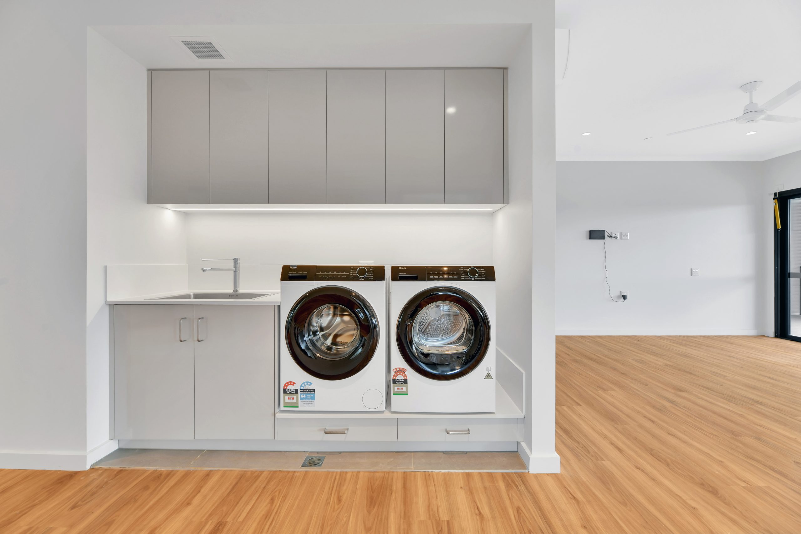 A modern laundry area with a front-loading washer and dryer, gray cabinets above and below, a small sink, and light wood flooring in a bright, open room.