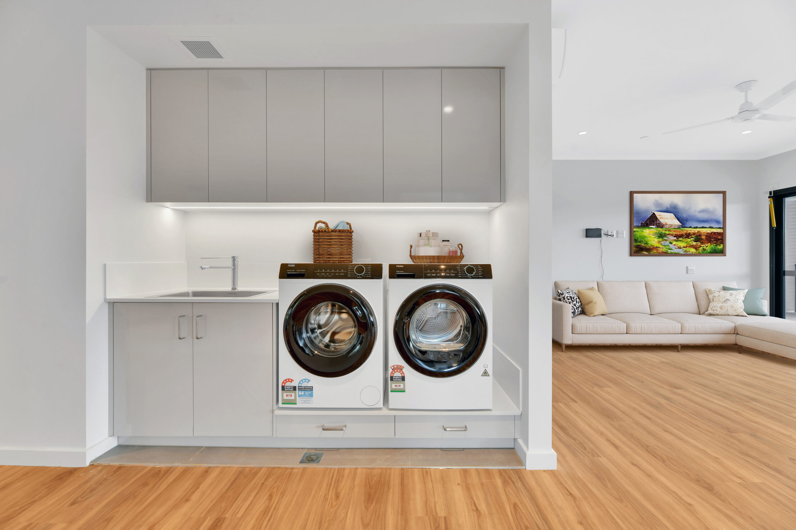 Modern laundry area with a washing machine and dryer, white cabinets, a countertop with a sink, and a wicker basket, adjacent to a bright living room with a beige sofa and wood flooring.