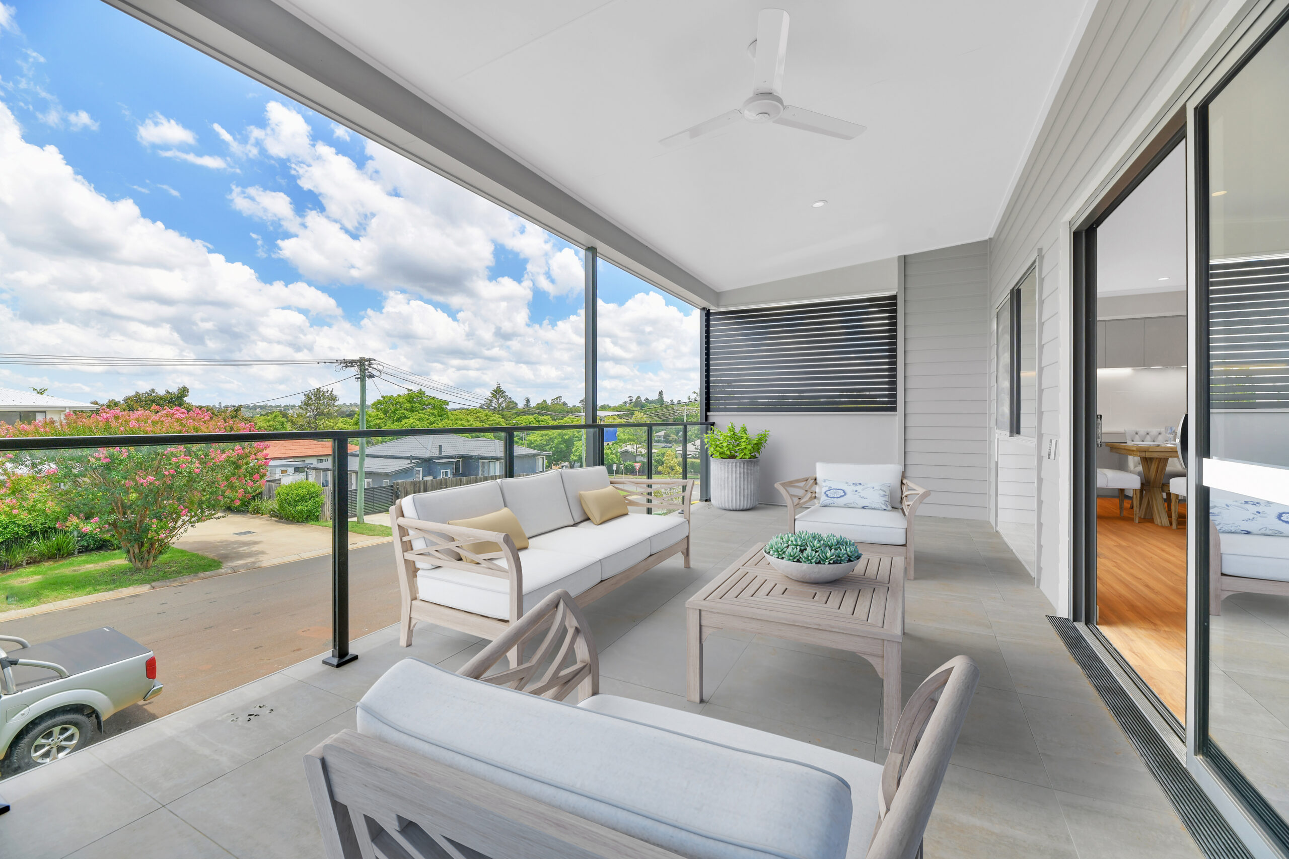 Modern covered balcony with white outdoor furniture, light wood tables, a potted plant, ceiling fan, and glass railing overlooking a suburban street with green trees and parked vehicles under a bright, partly cloudy sky.