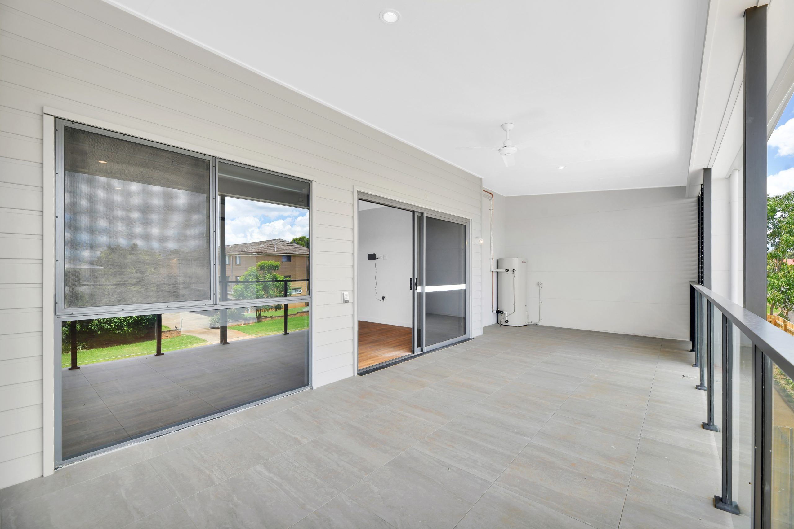 Spacious covered balcony with tiled floor, glass railing, sliding glass doors, and windows overlooking a green outdoor area. Ceiling fan and recessed lighting are visible above.