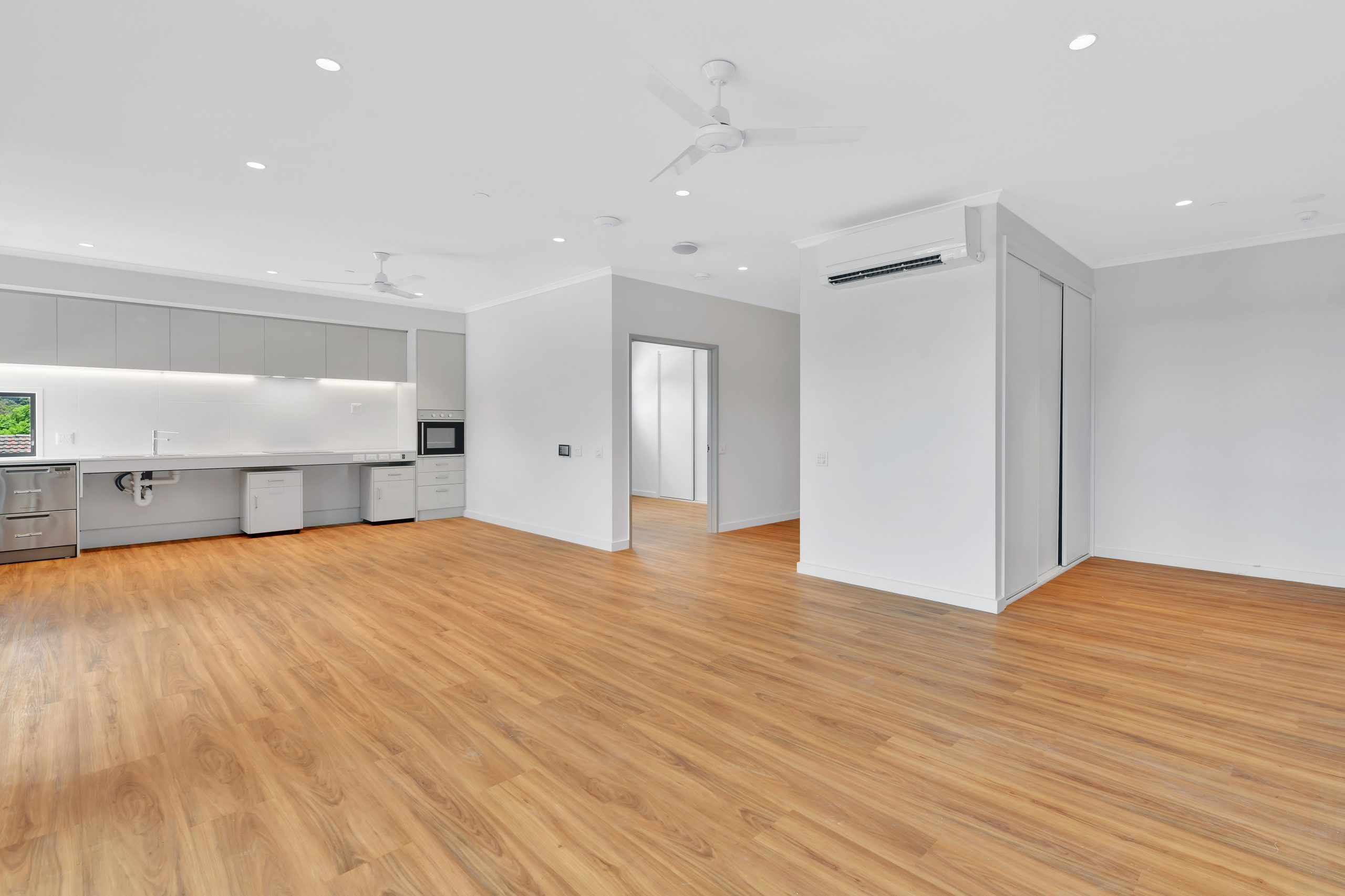 Spacious, modern open-plan room with light wood flooring, white walls, a kitchen area with white cabinets and appliances, ceiling fans, recessed lighting, and an air conditioning unit on the wall.