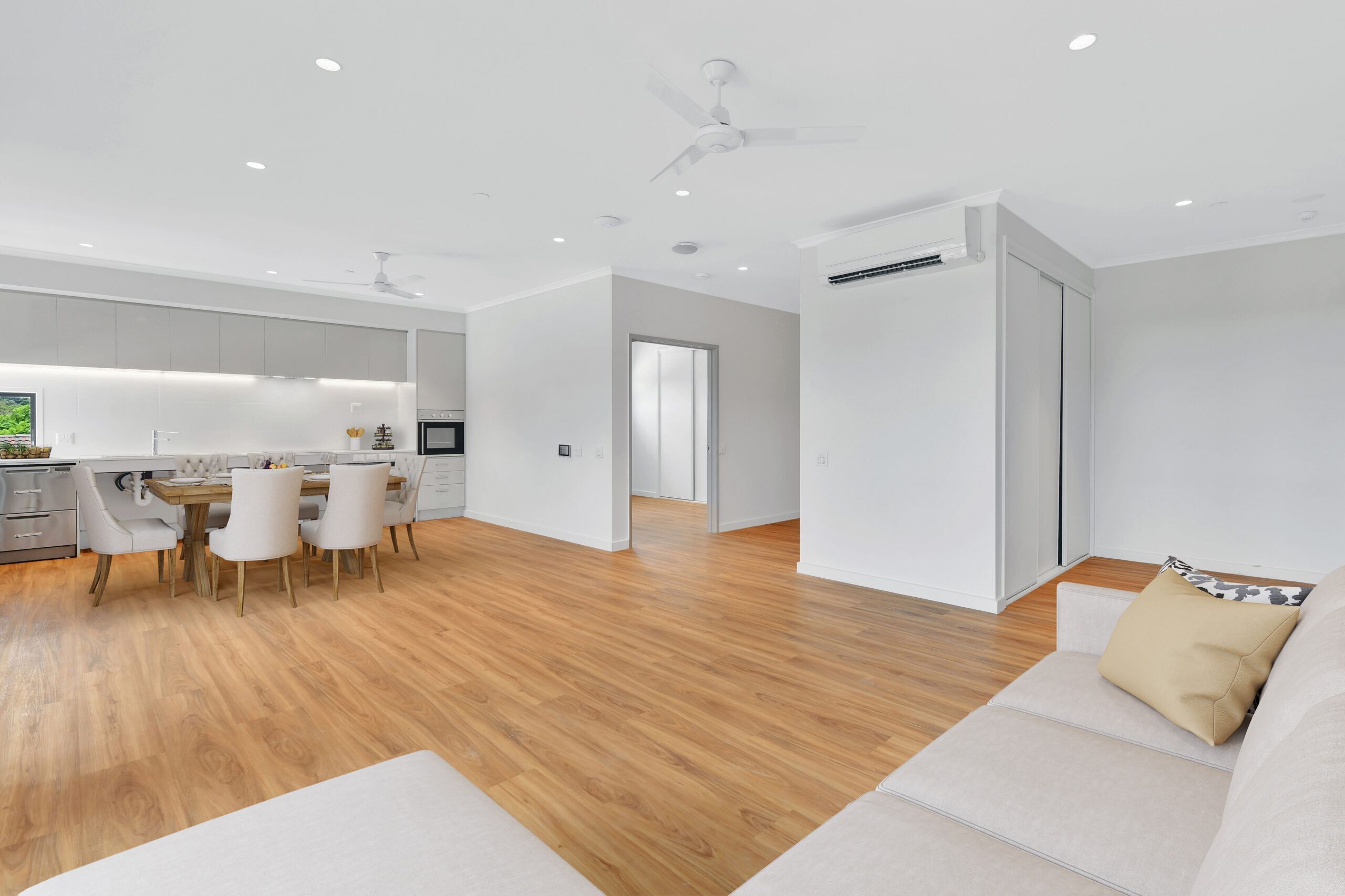A spacious, modern open-plan living area with light wood flooring, a beige sofa, a dining table with six chairs, a white kitchen with built-in appliances, and bright white walls and ceiling.