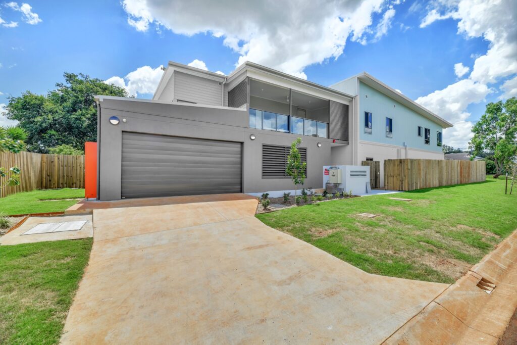 Modern two-story house with a gray exterior, large garage door, balcony, and a clean driveway, surrounded by green lawn and trees under a blue sky with clouds.