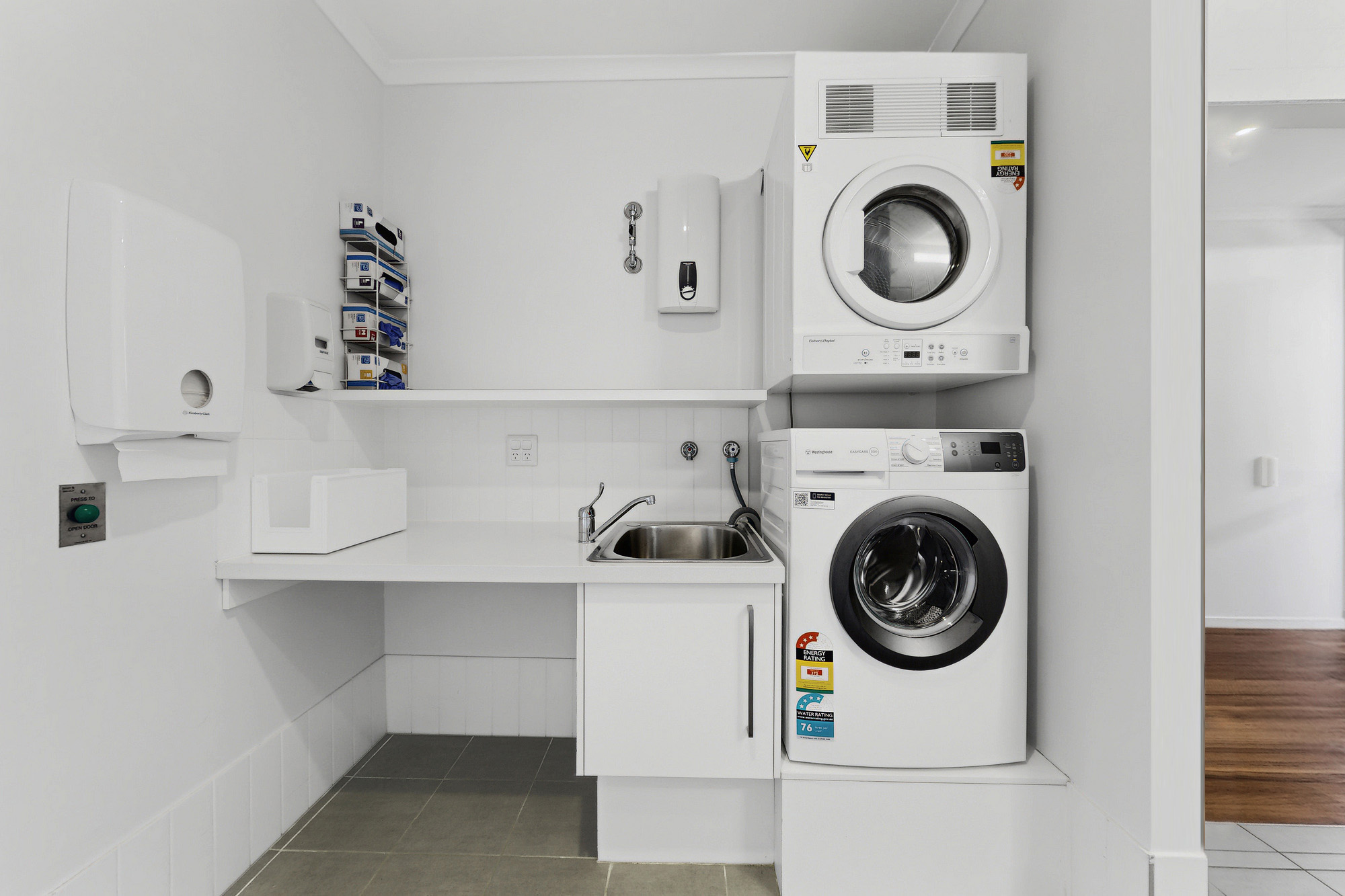 Modern laundry room with a stacked white dryer and washing machine, a small sink, white countertop, cleaning supplies on shelves, and tiled floor in a bright, minimalist setting.