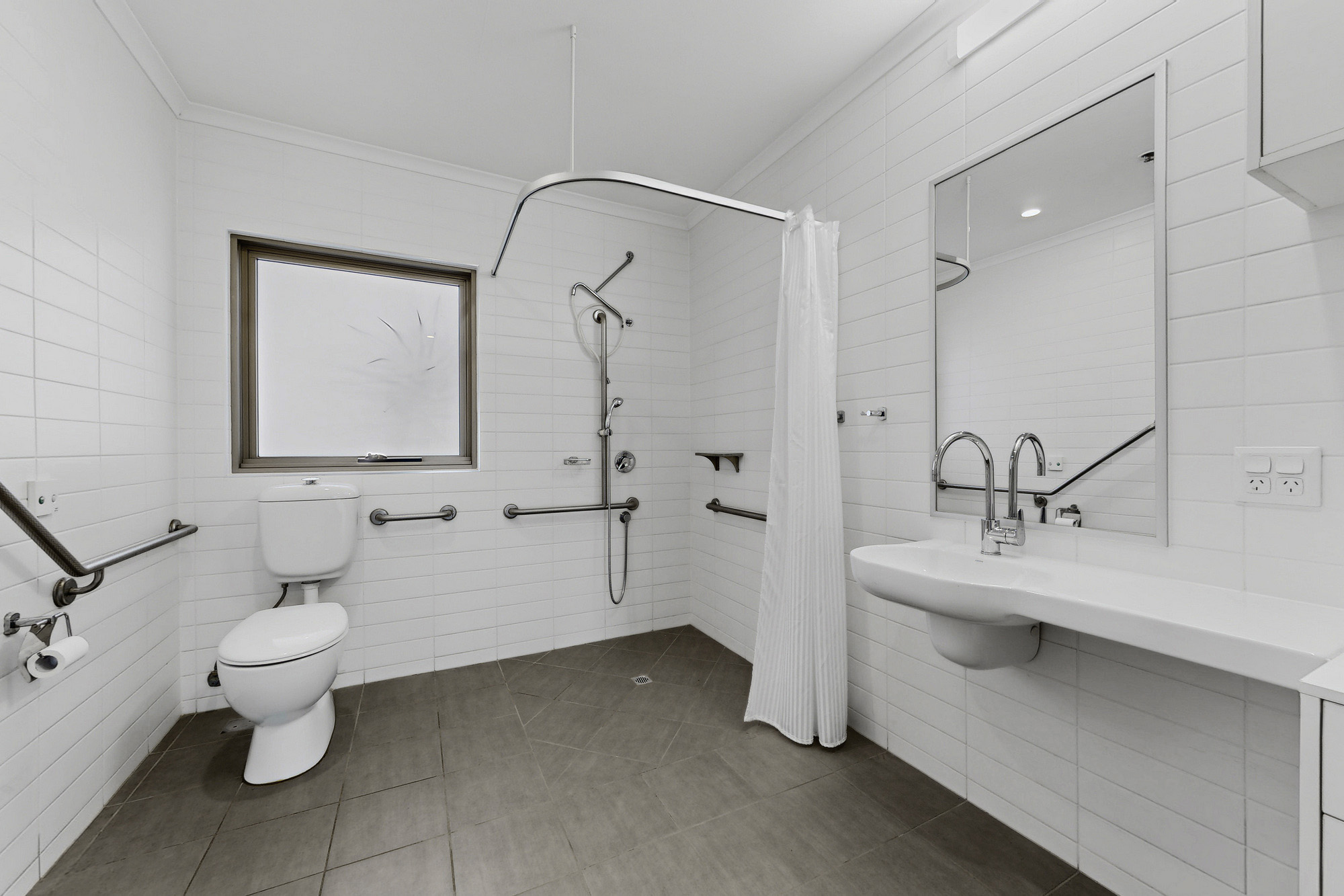 A modern, accessible bathroom with white tiles, grab bars, a wall-mounted toilet, a roll-in shower with a curtain, a handheld showerhead, and a large sink under a wide mirror.
