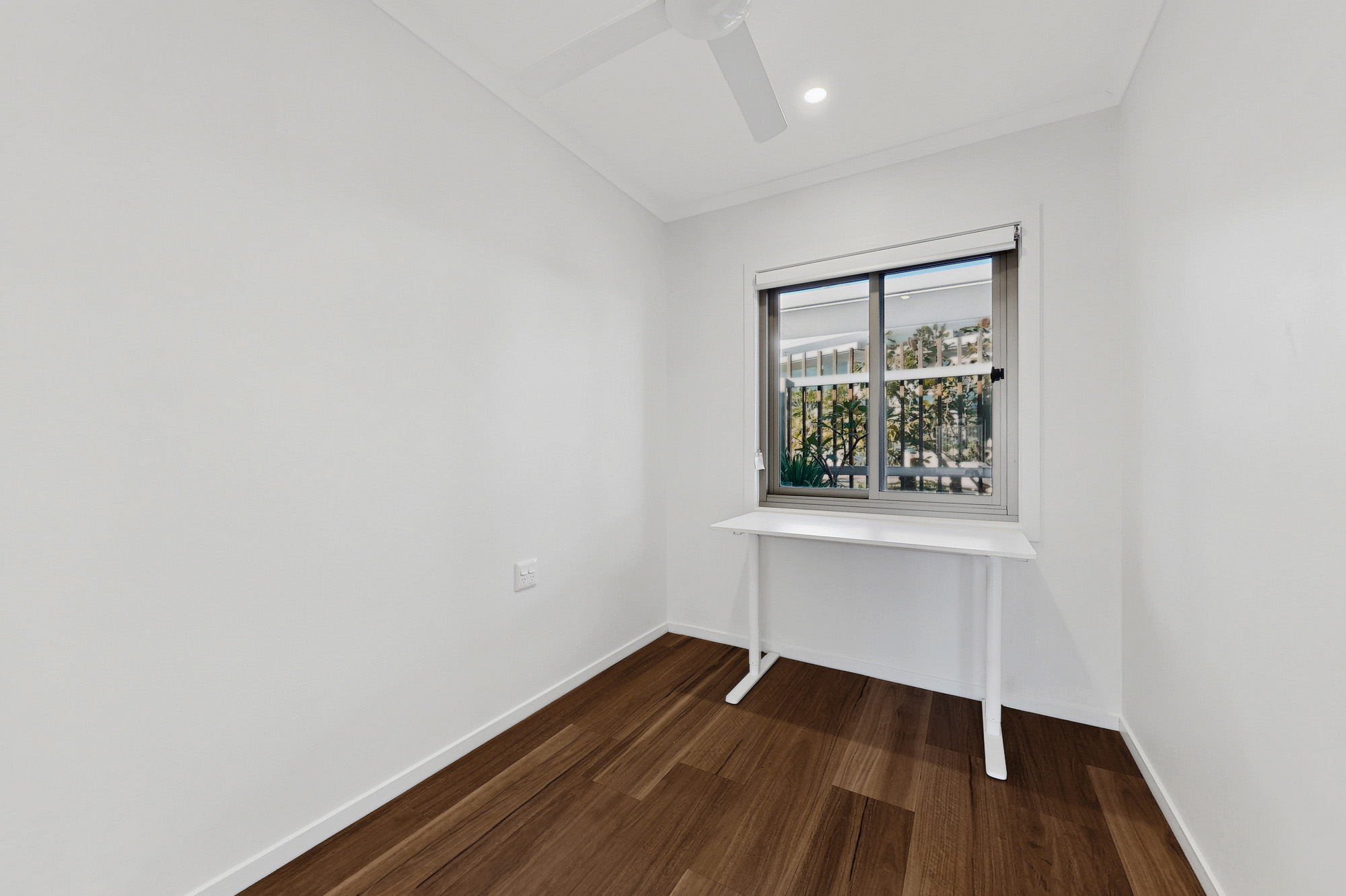 A small, bright room with white walls, a wooden floor, a ceiling fan, and a window. A simple white desk is placed under the window, and greenery is visible outside.