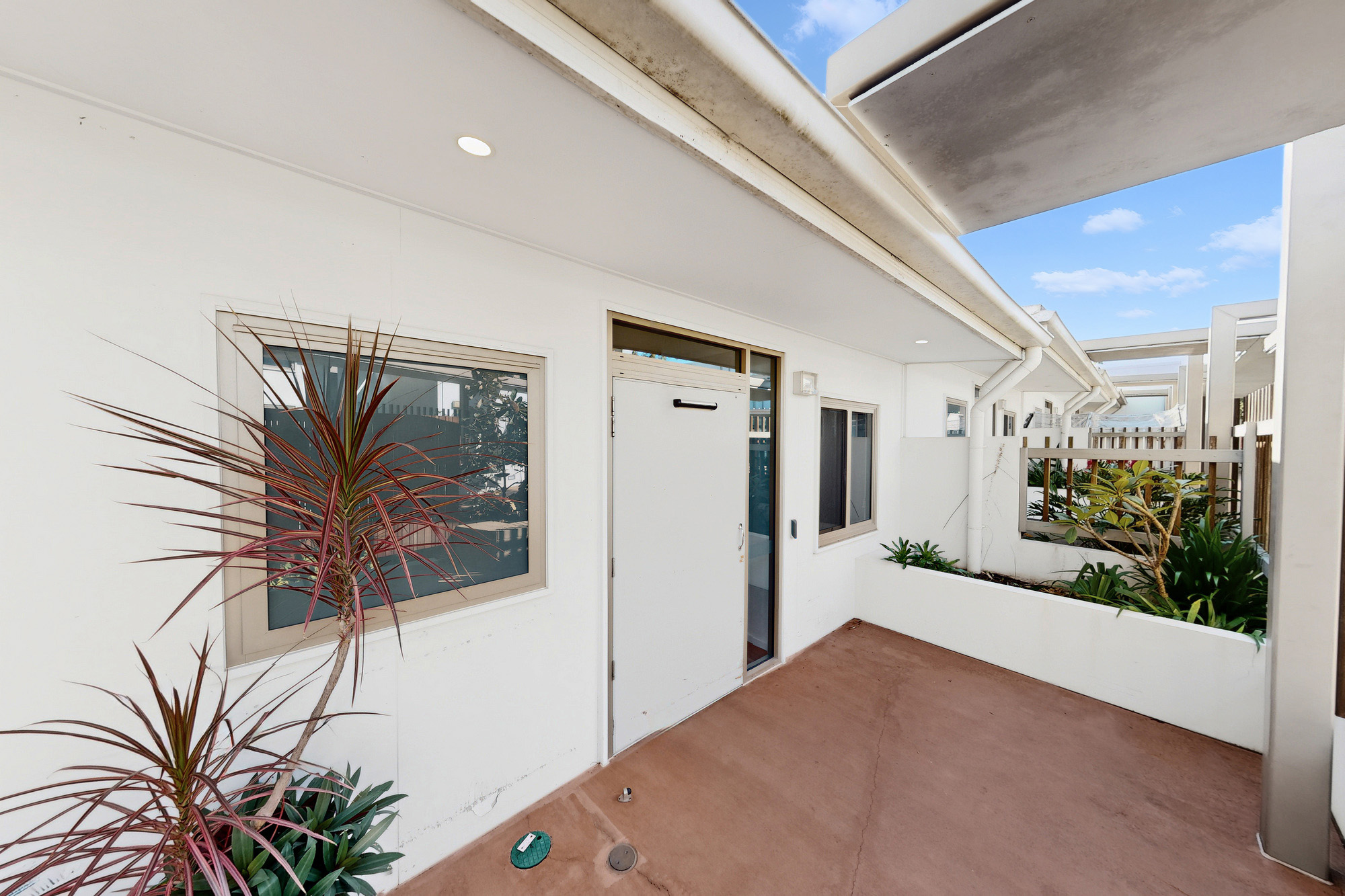 A small outdoor patio area with a reddish floor, white walls, potted plants, two windows, and a white door; the area is partially covered and bordered by a low wall with more plants.