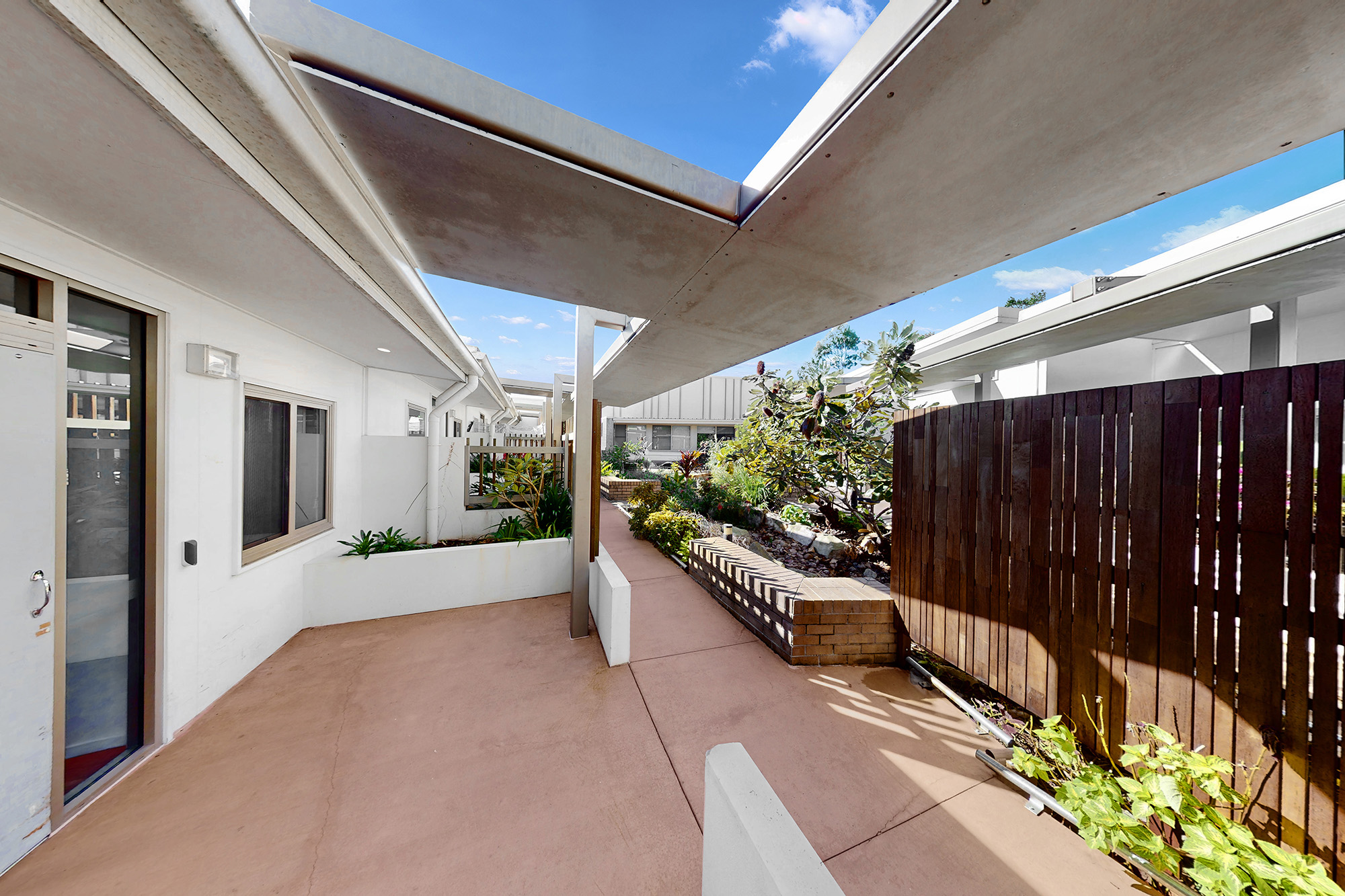 A modern outdoor patio with a covered walkway, potted plants, a wooden fence, and white buildings under a blue sky with scattered clouds.